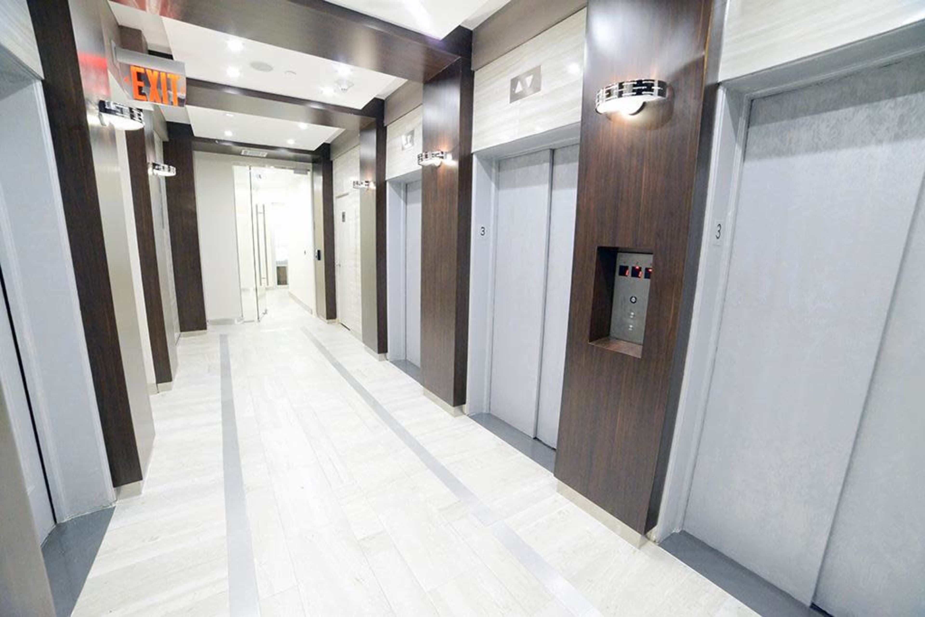 A modern hotel hallway with multiple elevator doors on either side, illuminated by wall sconces and featuring a visible exit sign at the end.