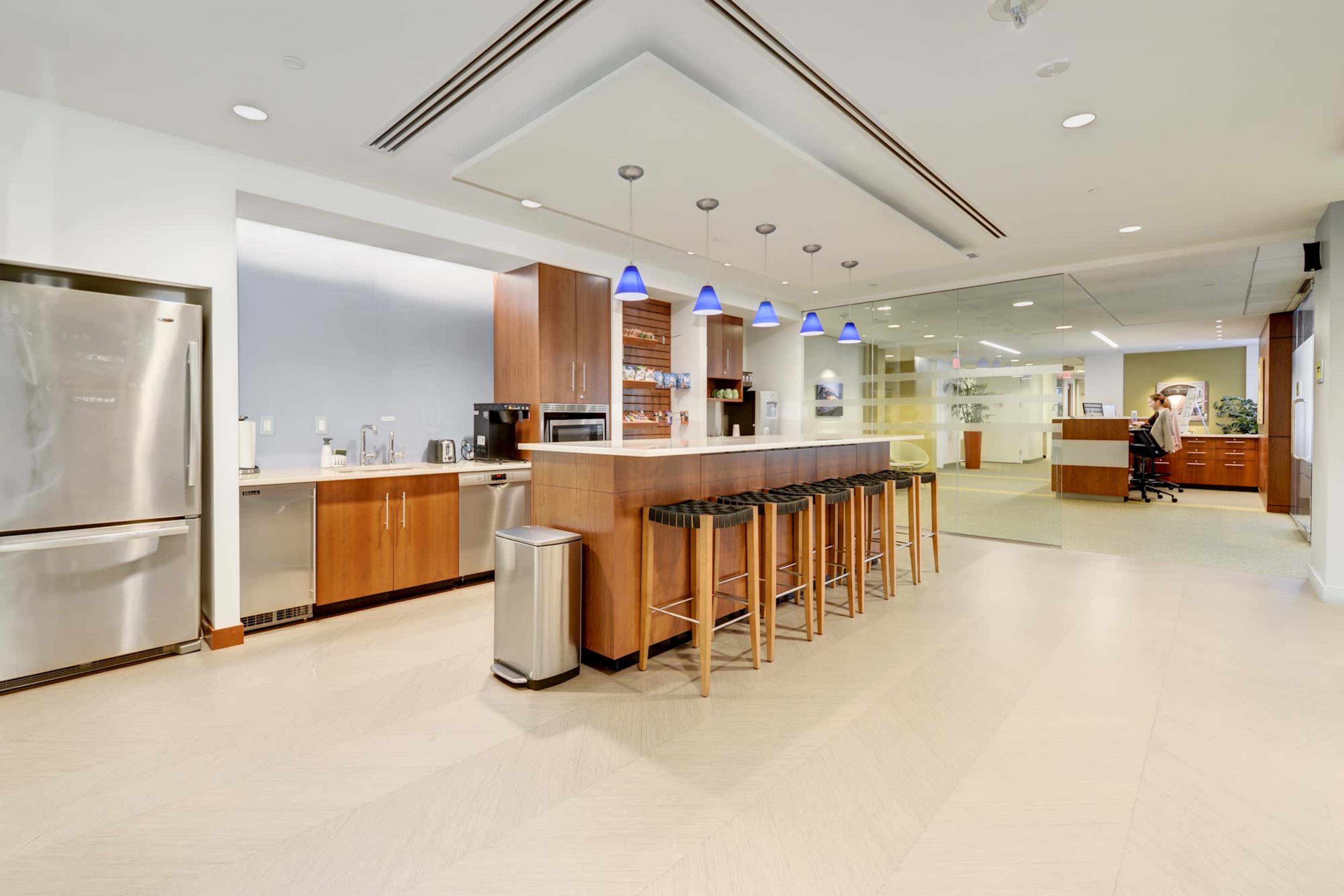 A modern kitchen area with wooden cabinets, bar stools, and stainless steel appliances in an office setting.