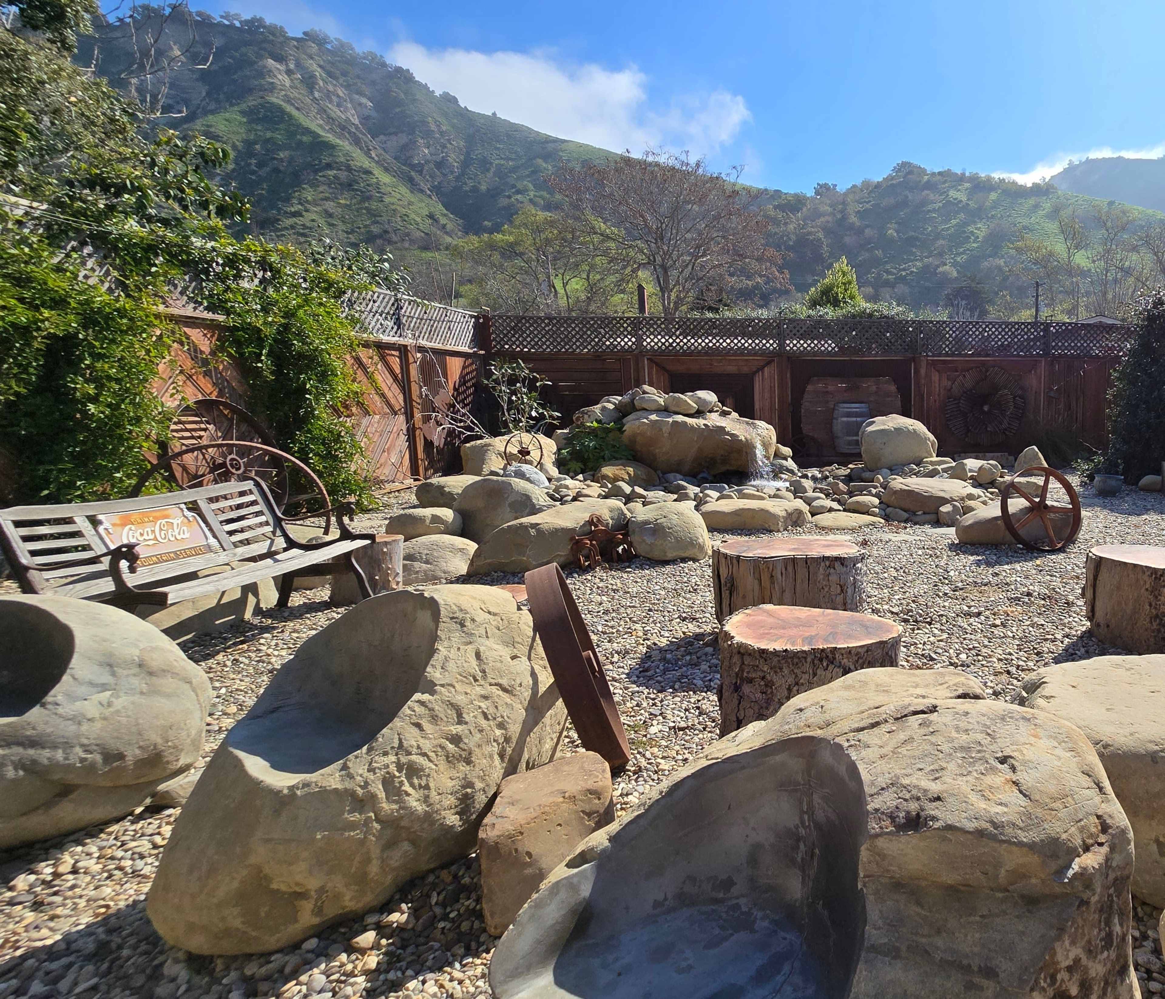A rock garden with large stones, wooden logs, and a bench, surrounded by a wooden fence and greenery under a clear blue sky.