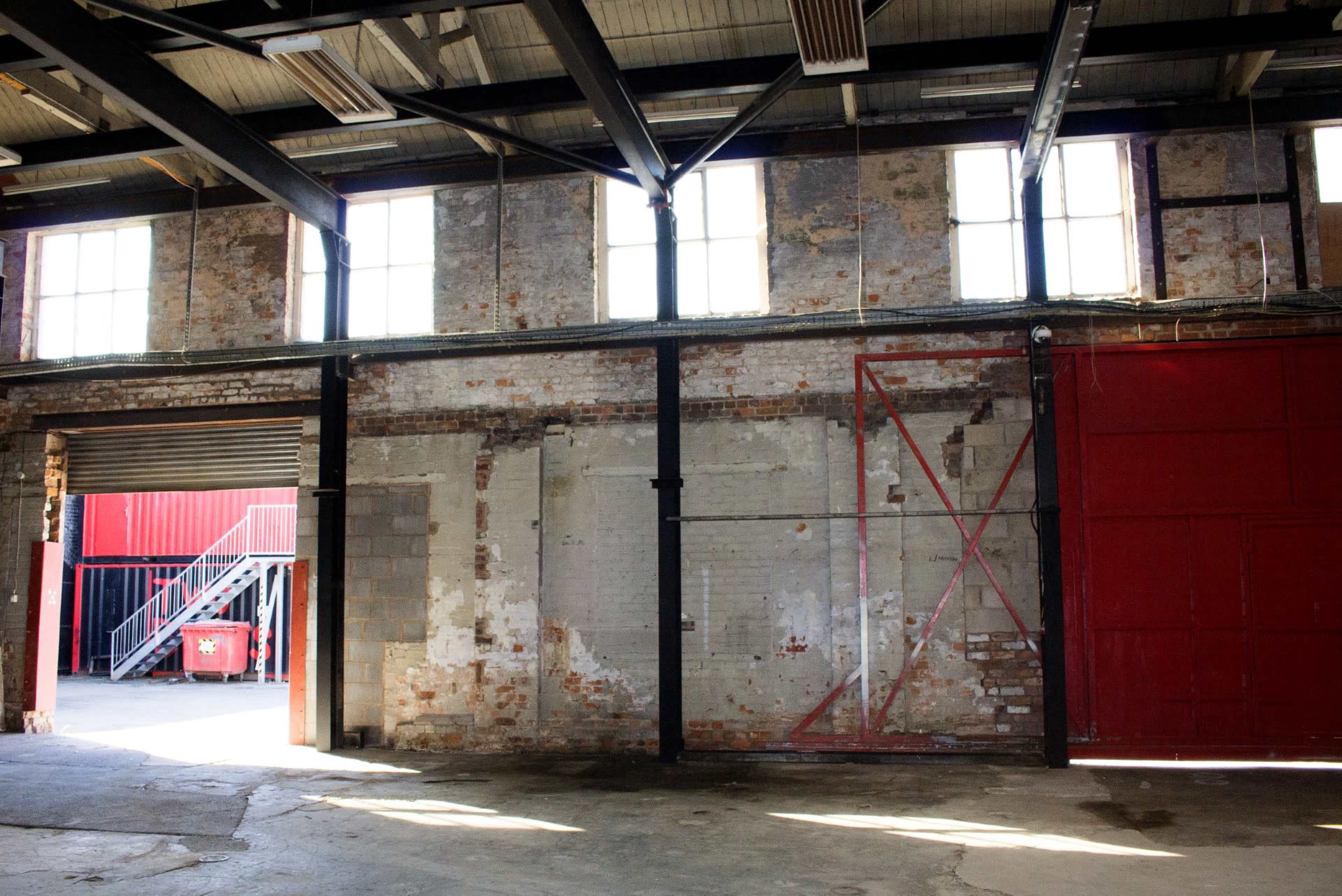 An interior view of a warehouse with exposed brick walls and large windows, featuring a red door and staircase visible in the background.