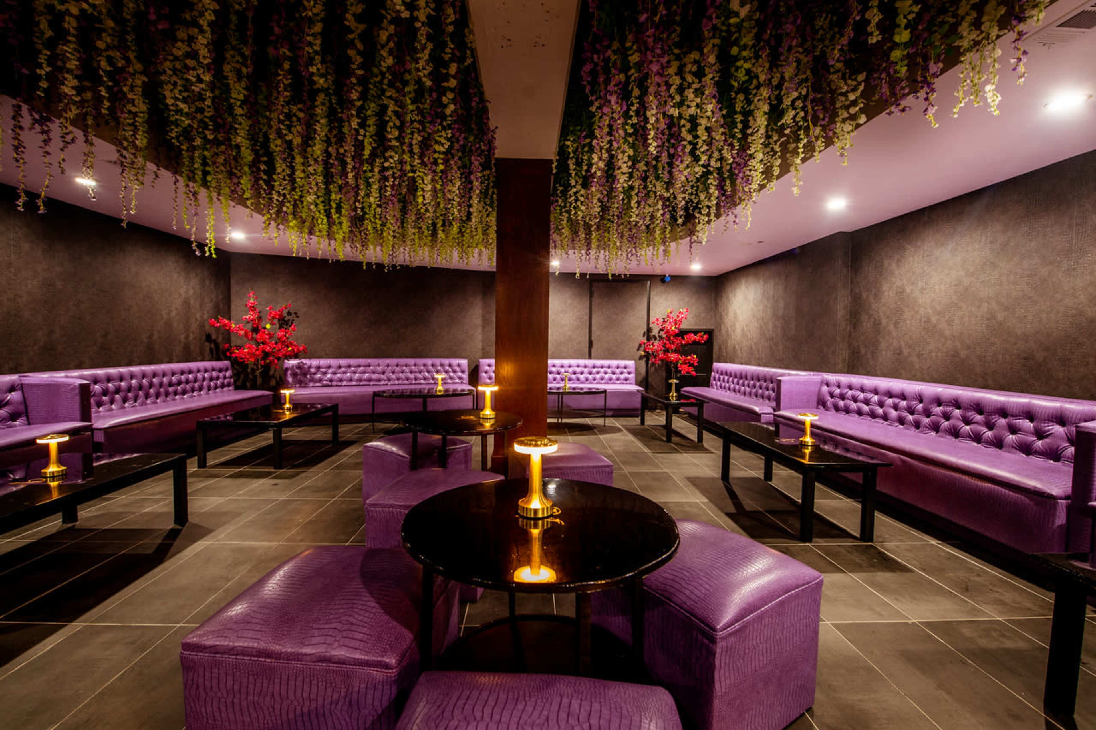 The scene features a dimly lit lounge with purple tufted seating, black tables, and decorative floral arrangements hanging from the ceiling.