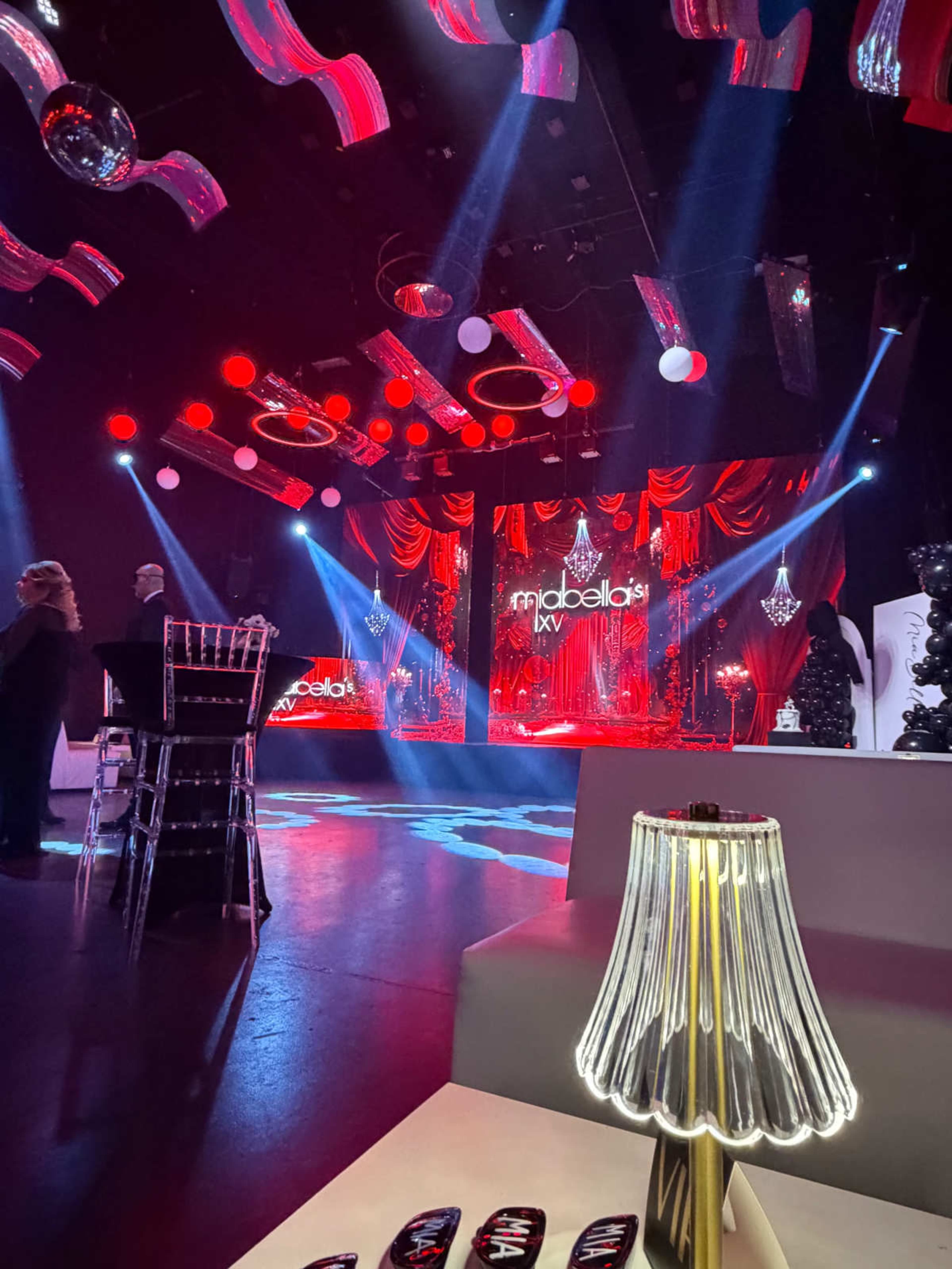The image shows a stage set up for an event, featuring red lighting, decorative elements, and chairs in the foreground.