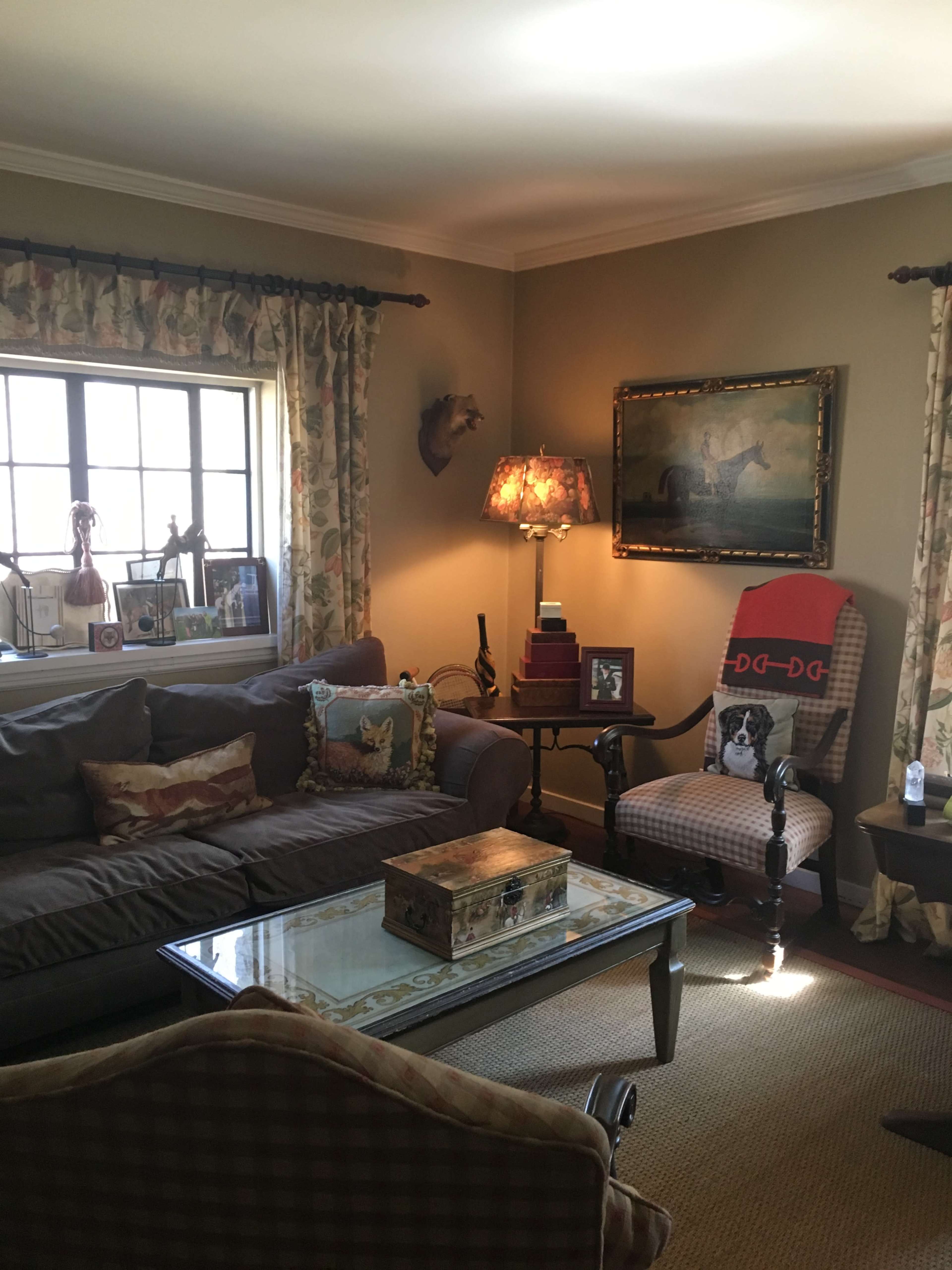 The image shows a cozy living room with a brown couch, a glass coffee table, and a patterned armchair beside a window adorned with floral curtains.