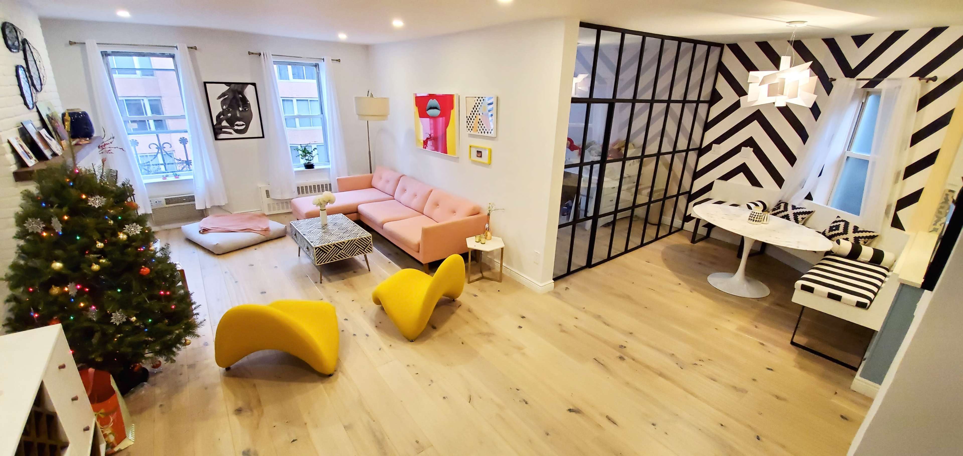 A modern, open-concept living space featuring a pink sofa, yellow chairs, a Christmas tree, and a dining area with black and white accents.