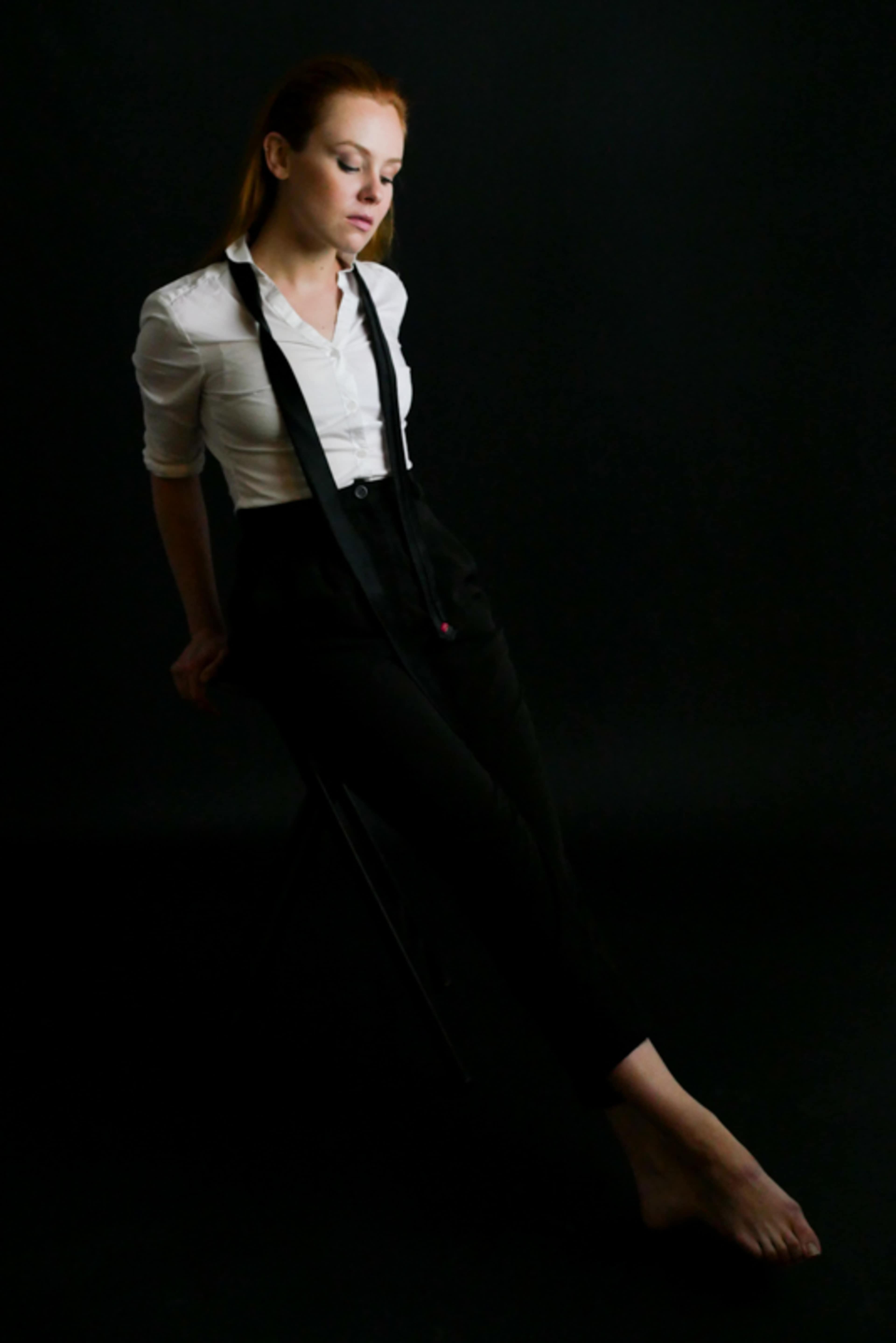 A woman in a white shirt and black pants sits gracefully on a stool against a dark backdrop.
