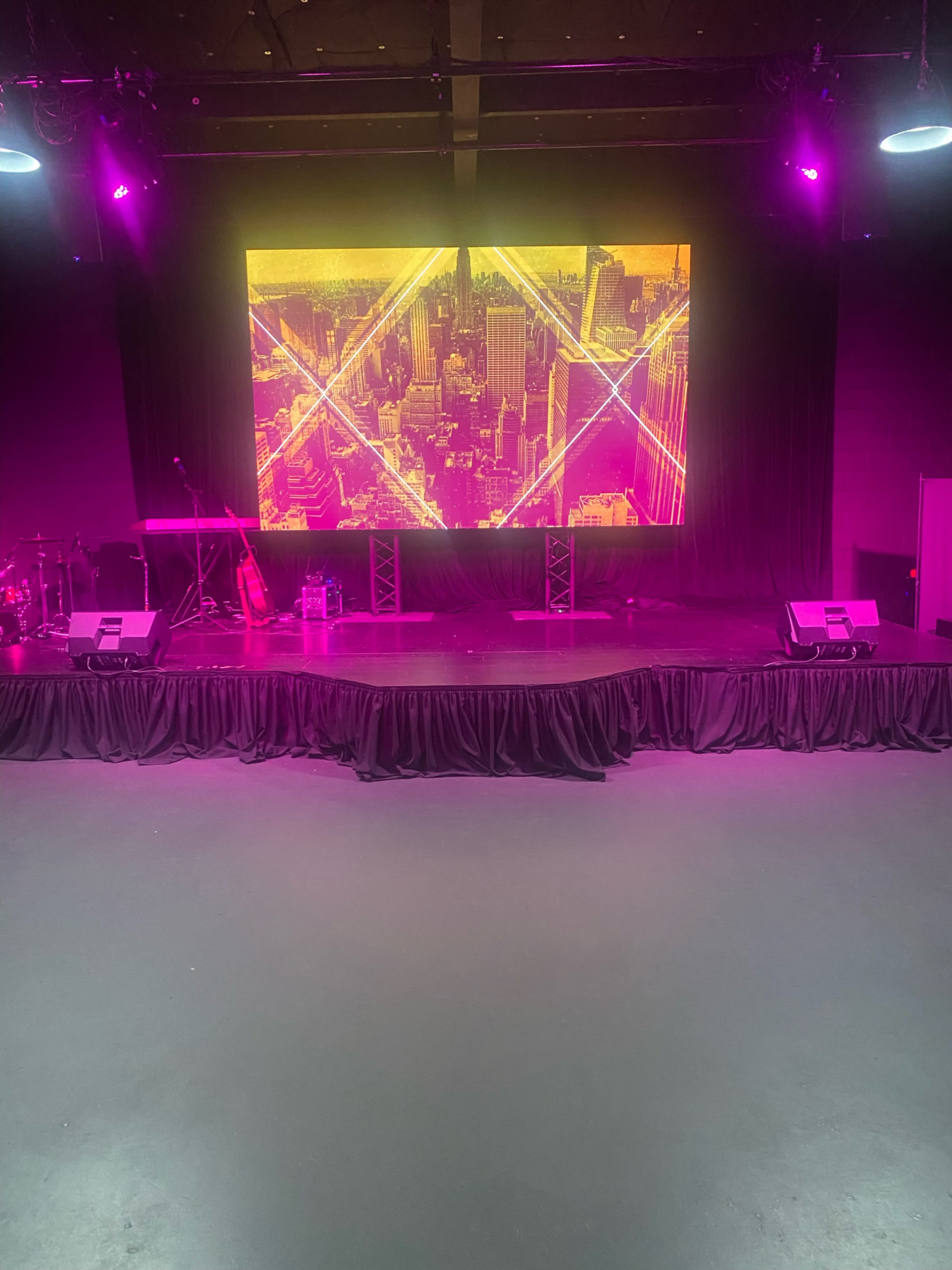 The image shows a stage setup with a large digital screen displaying a cityscape, flanked by musical instruments and an illuminated backdrop in a darkened venue.