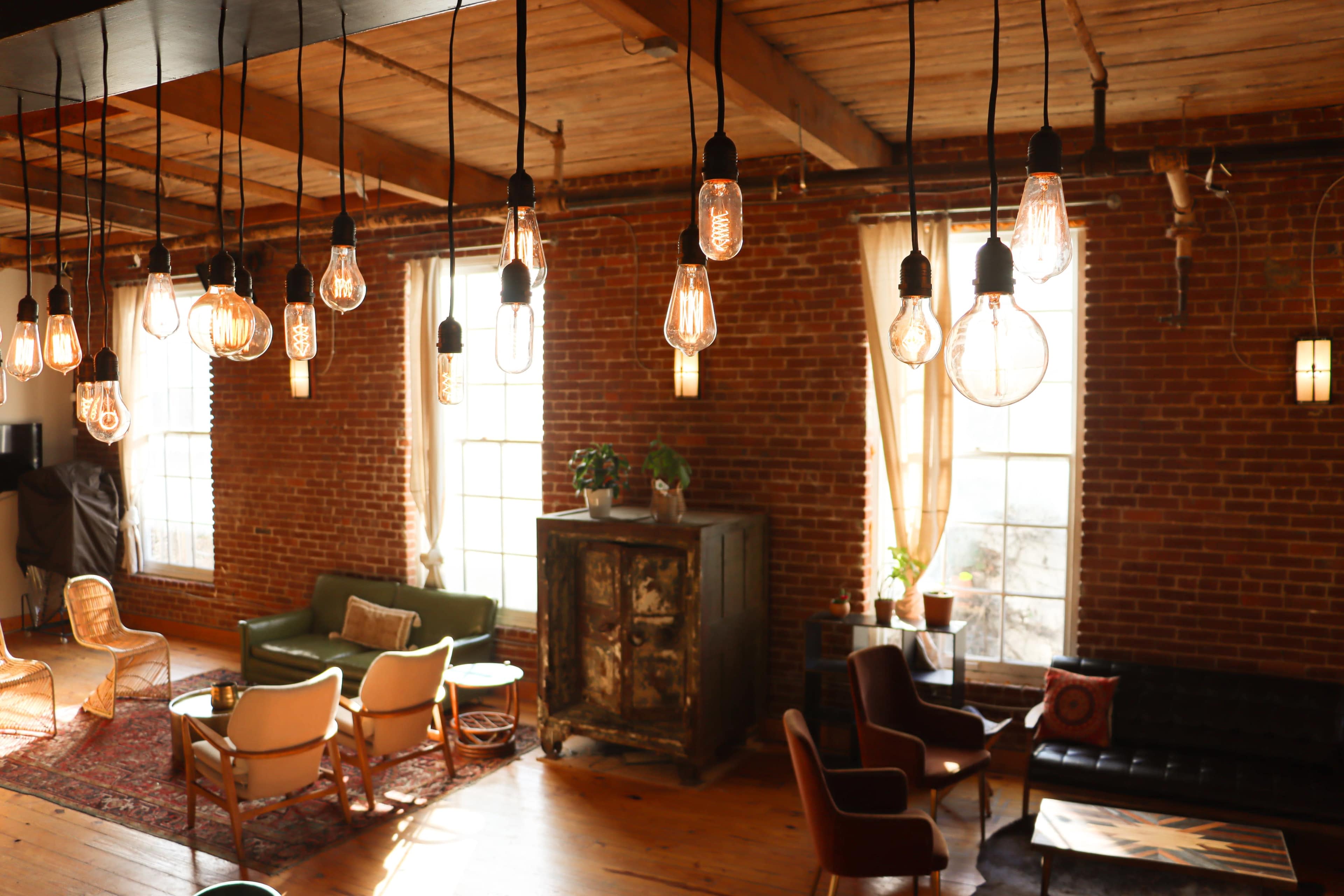 A spacious interior features exposed brick walls, large windows, and various seating areas illuminated by hanging light bulbs.