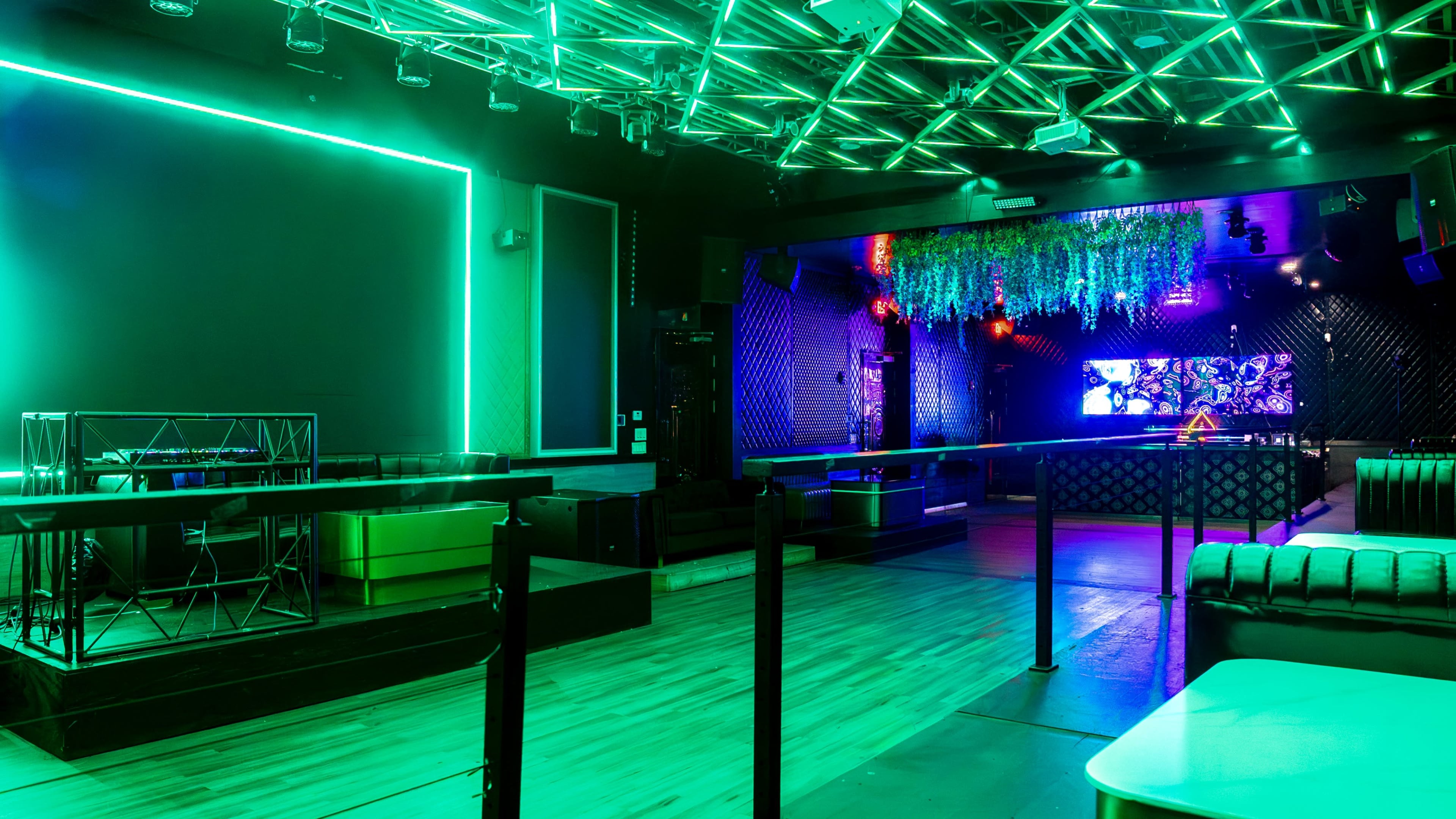 The image shows an empty nightclub interior featuring vibrant green lighting, modern seating arrangements, and a DJ stage with a colorful backdrop.