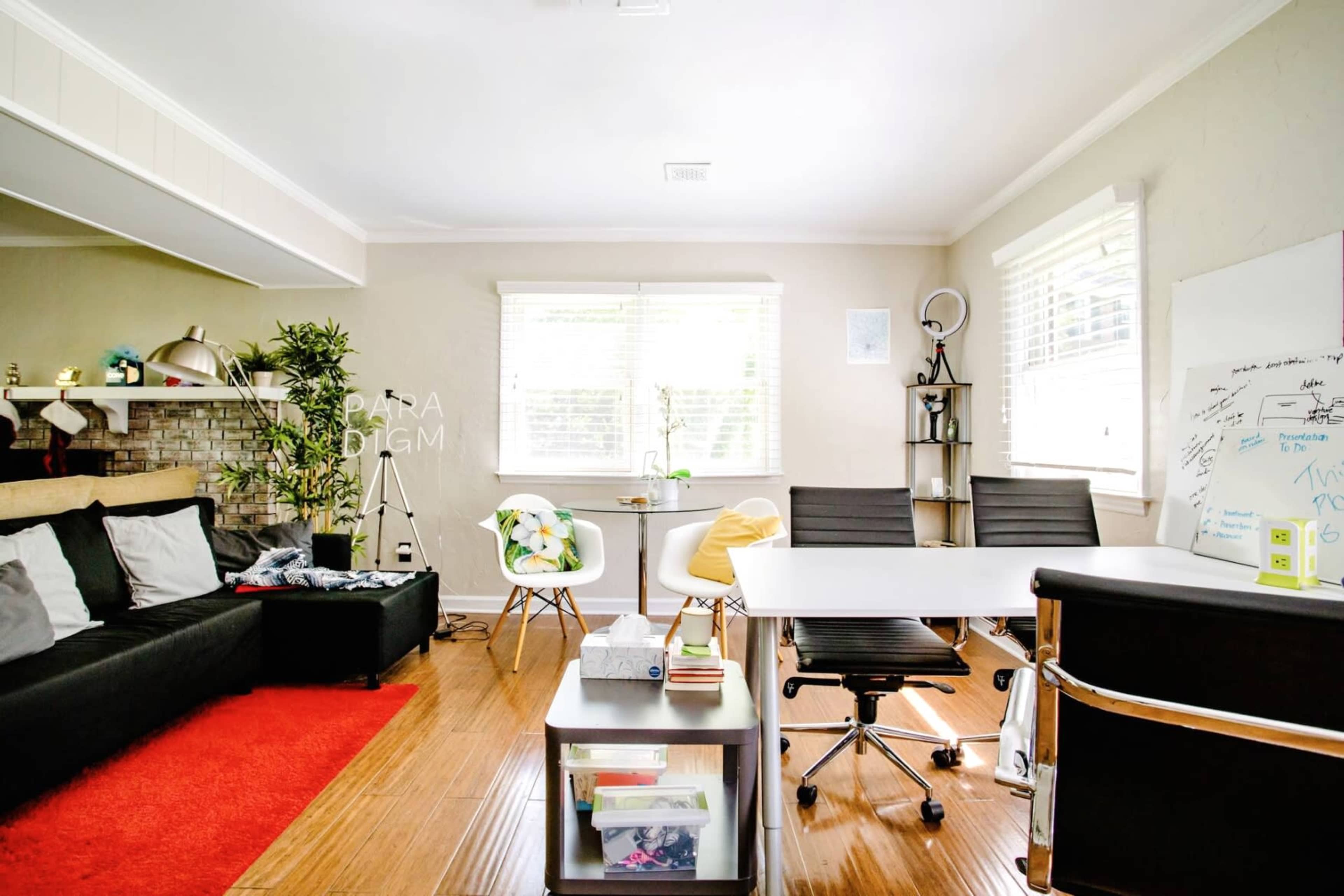 The image shows a bright, modern office space with a large white desk, comfortable seating, and large windows allowing natural light to enter.