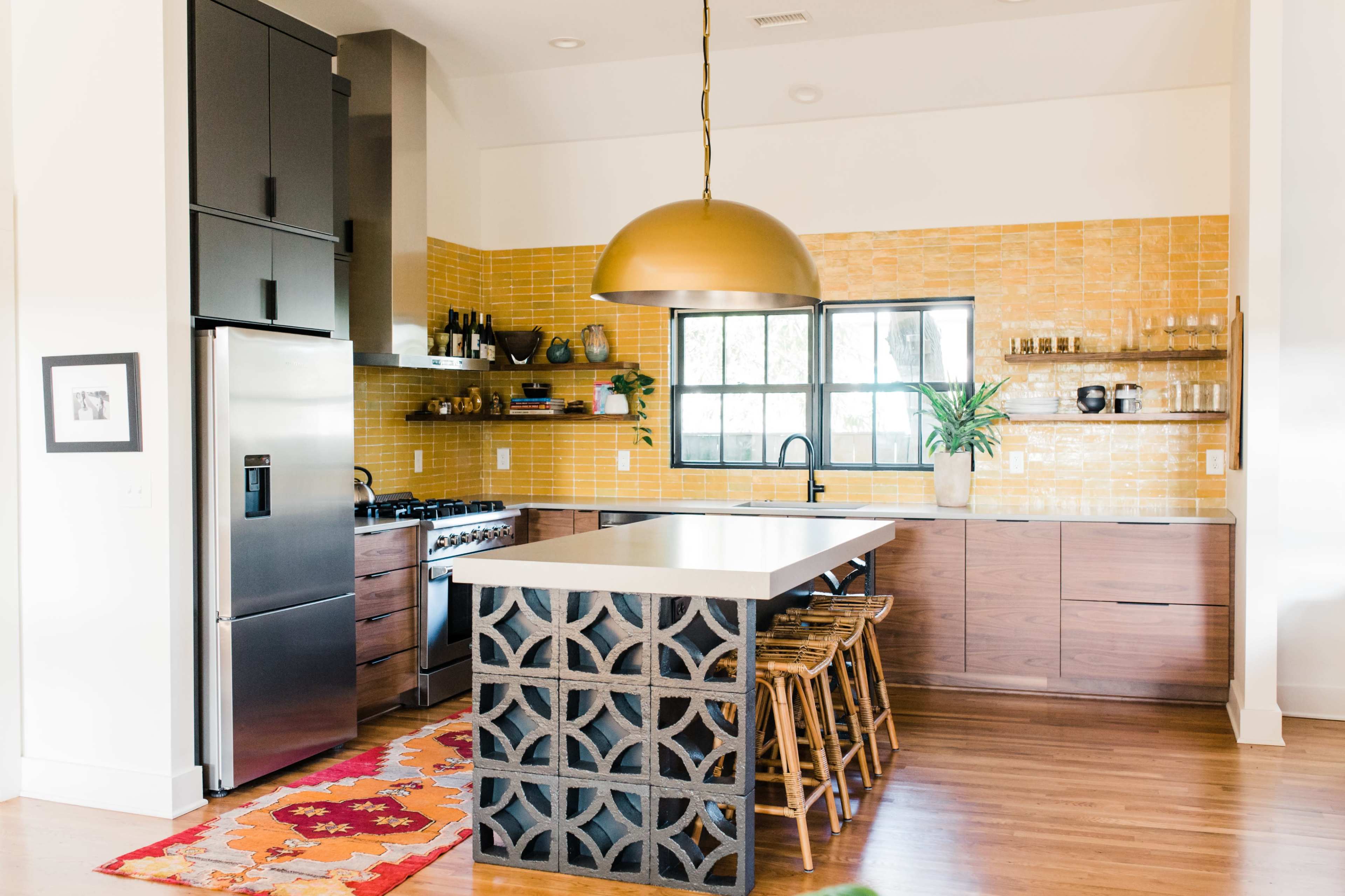 A modern kitchen features a large central island with wooden stools, a stainless steel refrigerator, and a striking yellow tiled backsplash.