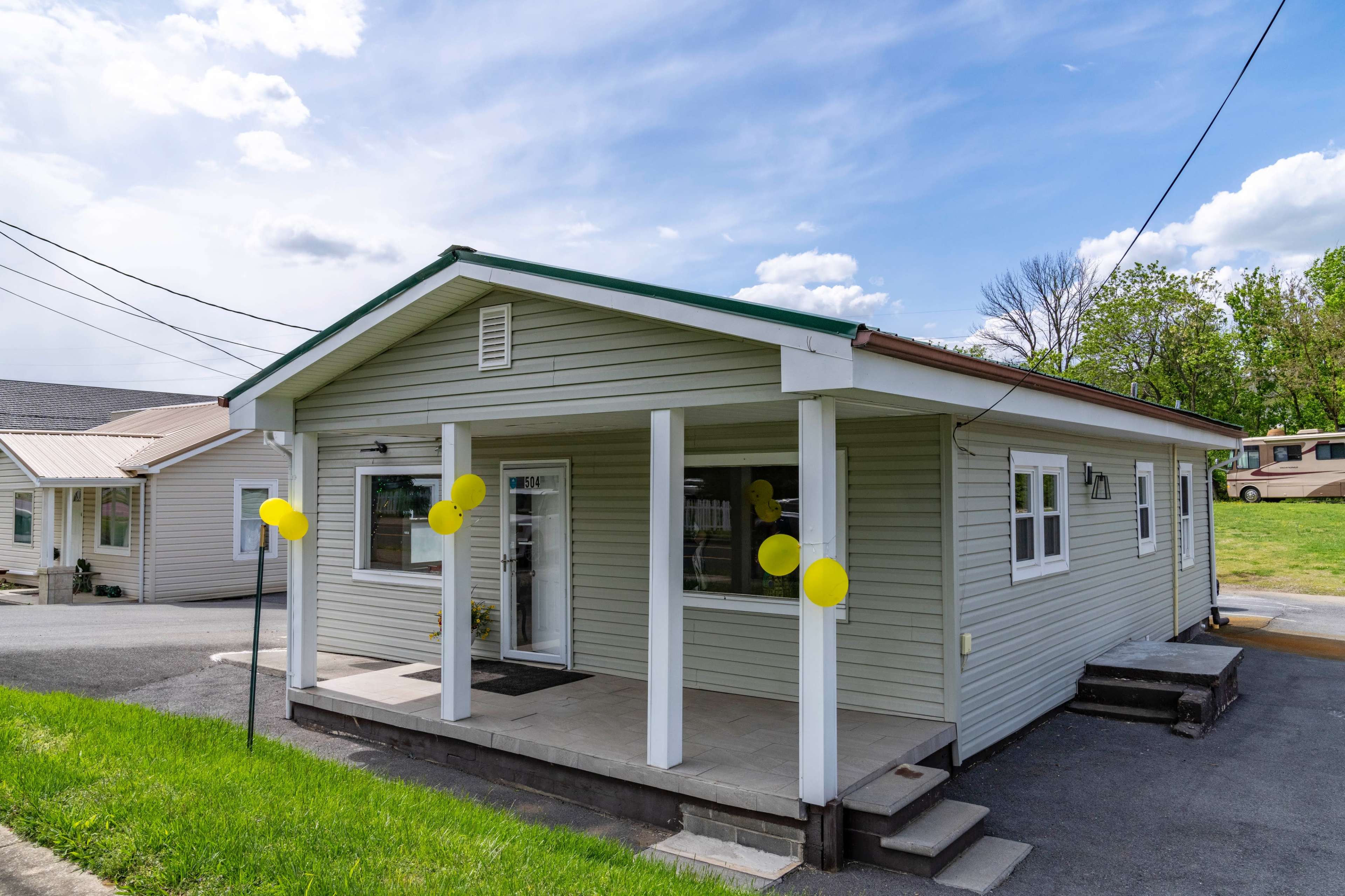 A single-story house with a green roof and yellow balloons on the front porch is situated on a grassy lot.