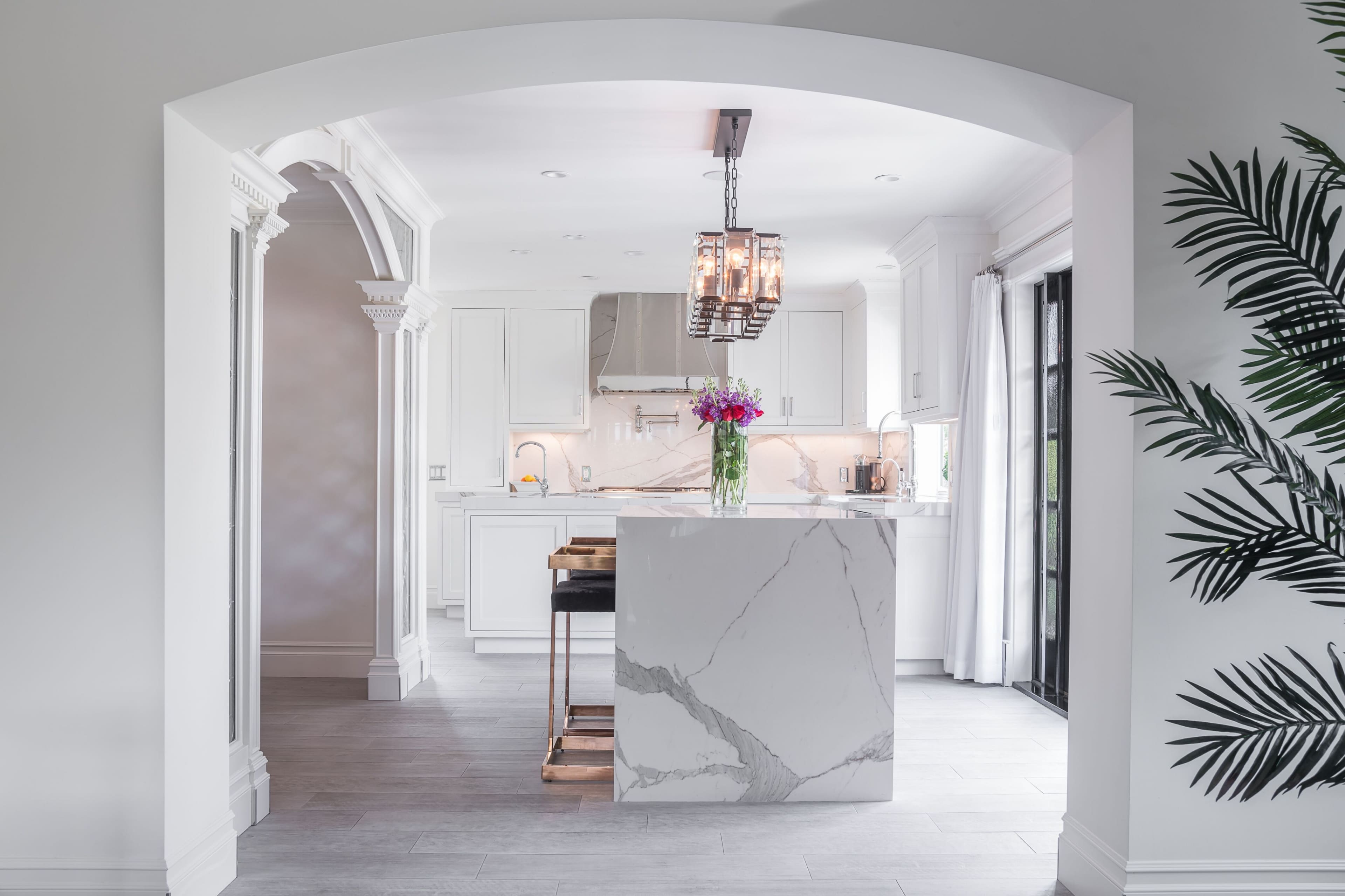 A modern kitchen features a large marble island with a bouquet of flowers, stainless steel appliances, and white cabinetry, viewed through an arched entryway.