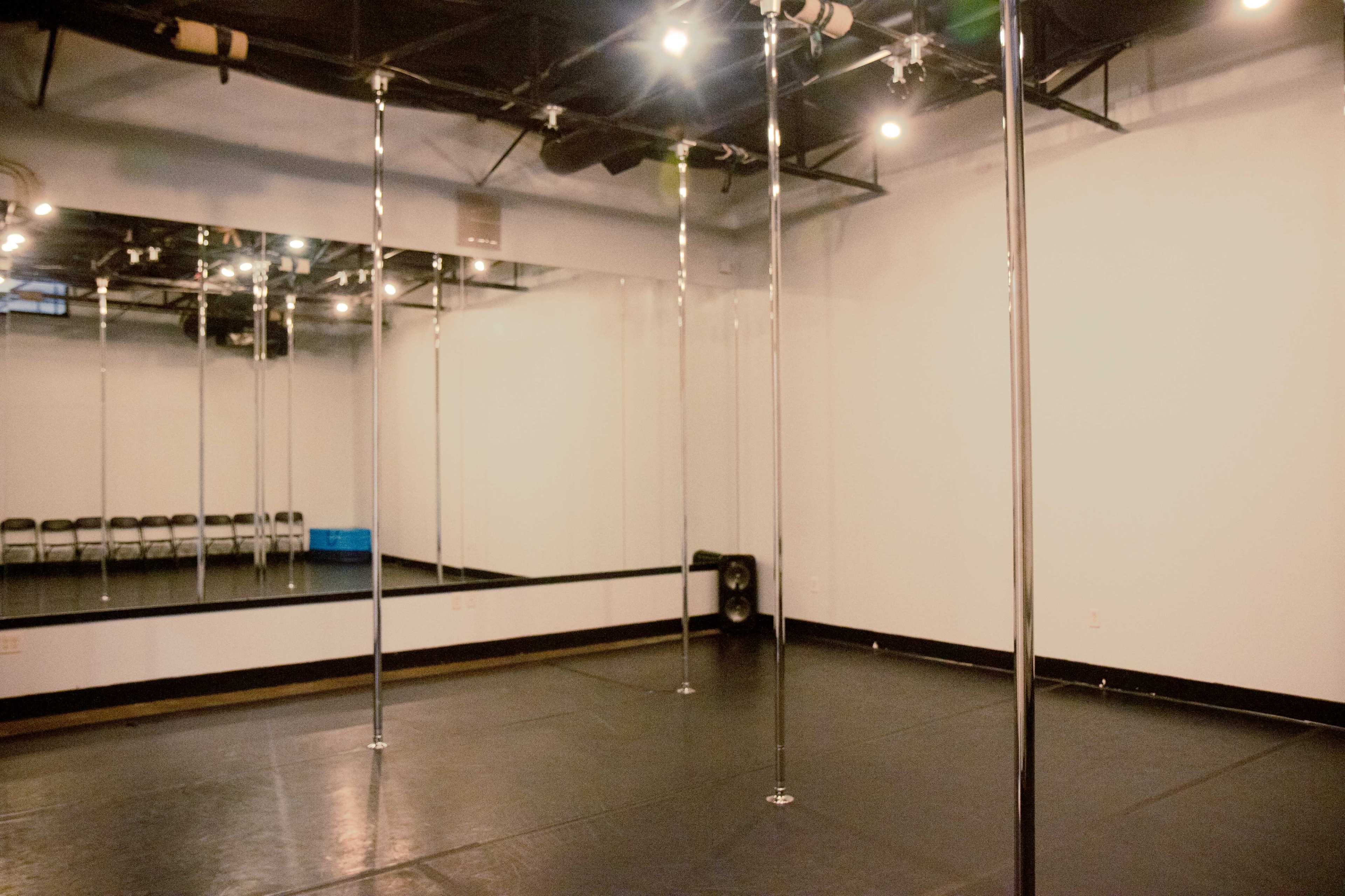 The image shows a studio with several polished poles, a large mirror, and a black floor, illuminated by overhead lights.