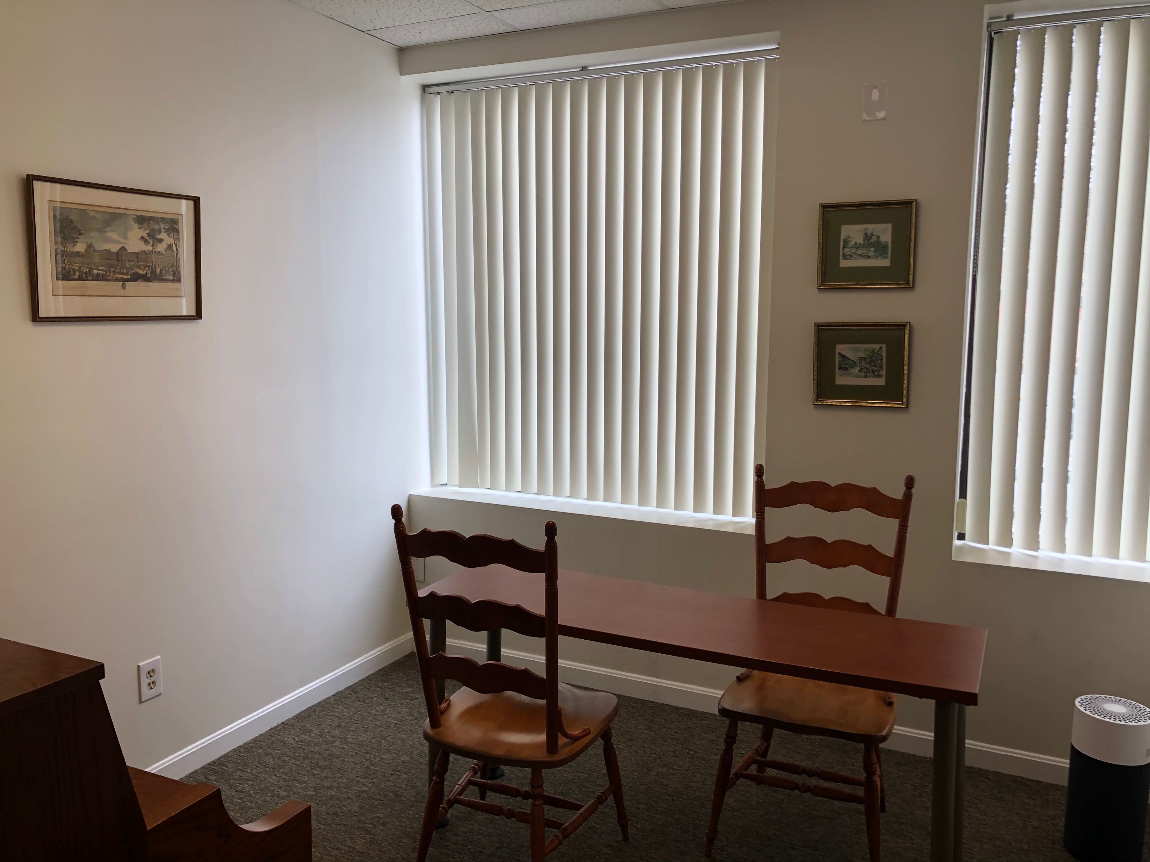 A small office space with two wooden chairs and a table positioned near large windows with vertical blinds.
