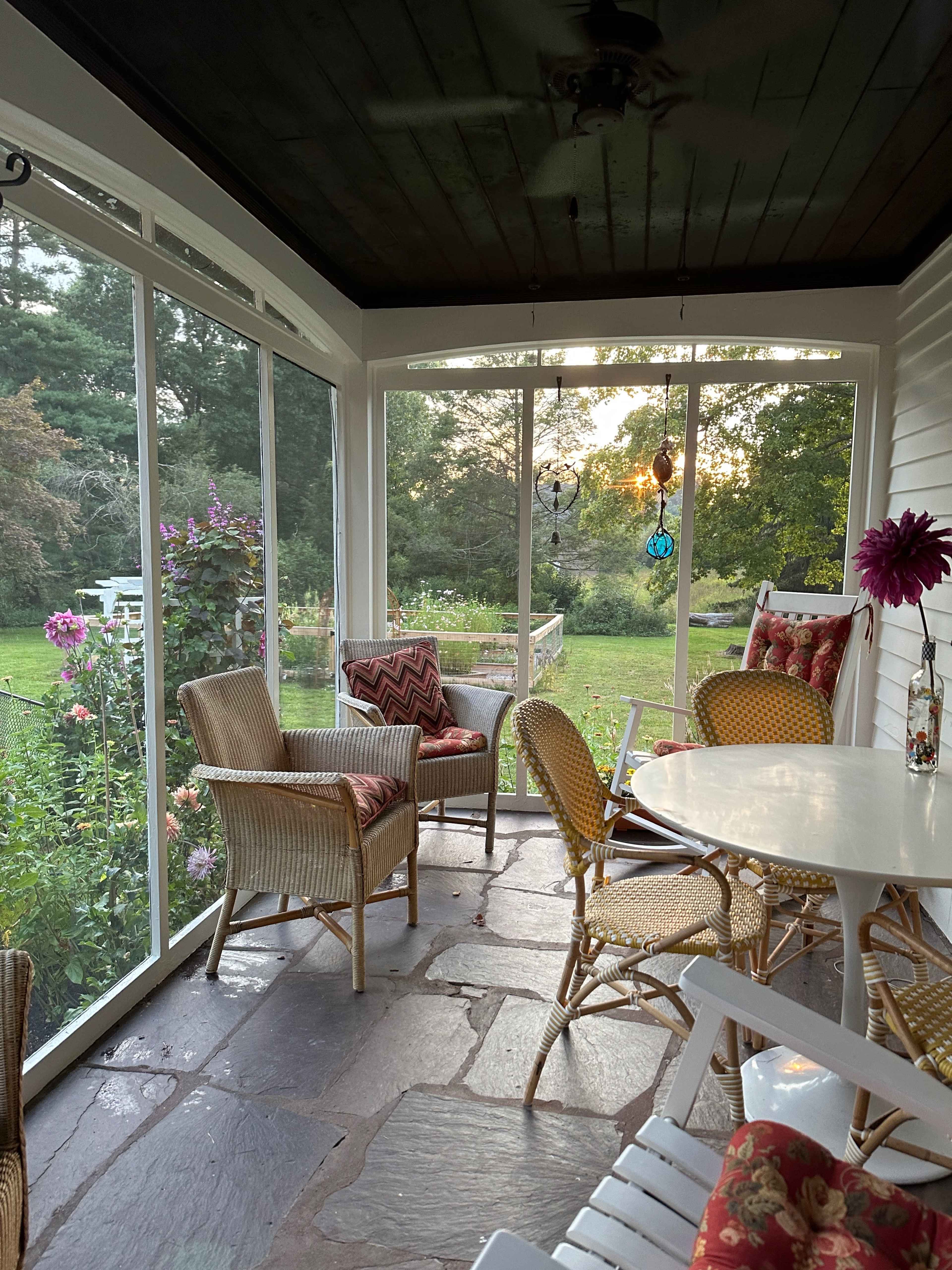 A screened porch with wicker chairs and a small table, surrounded by greenery.