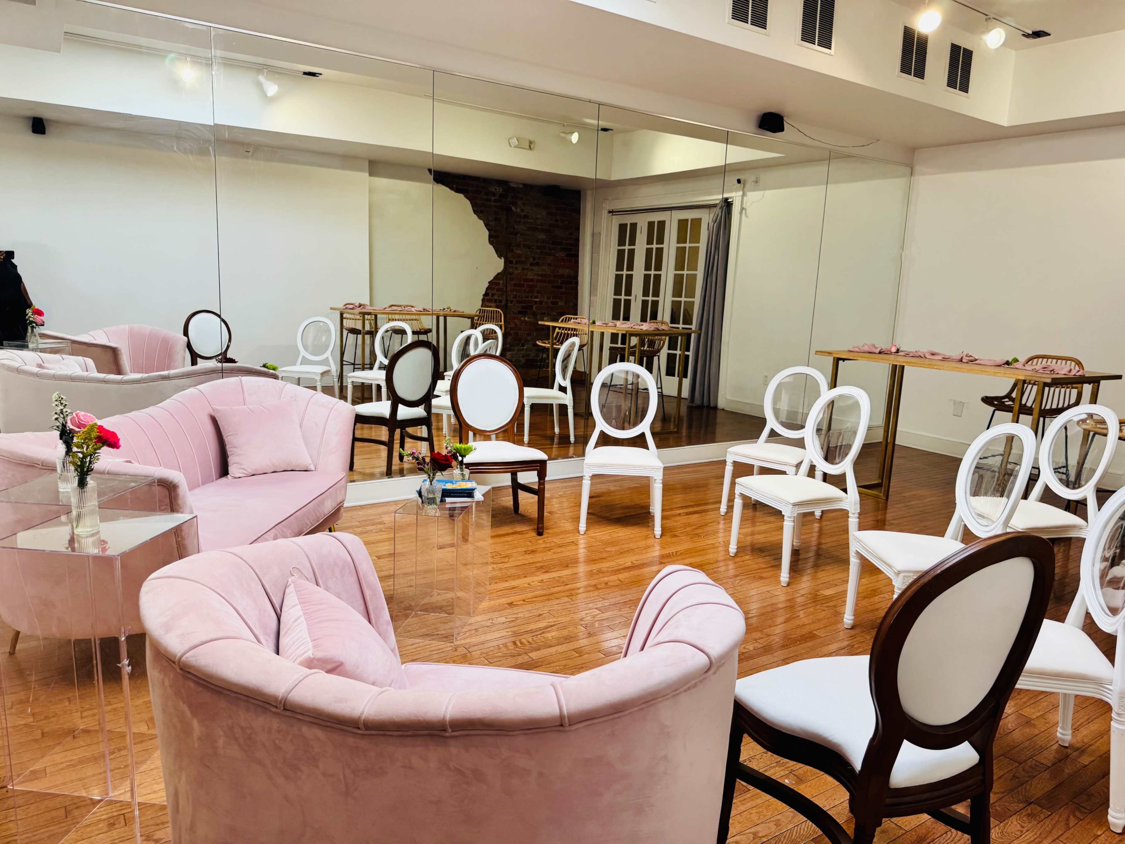 A spacious, modern room features pink velvet sofas and white chairs arranged around a table, with mirrors and wooden floors enhancing the ambiance.