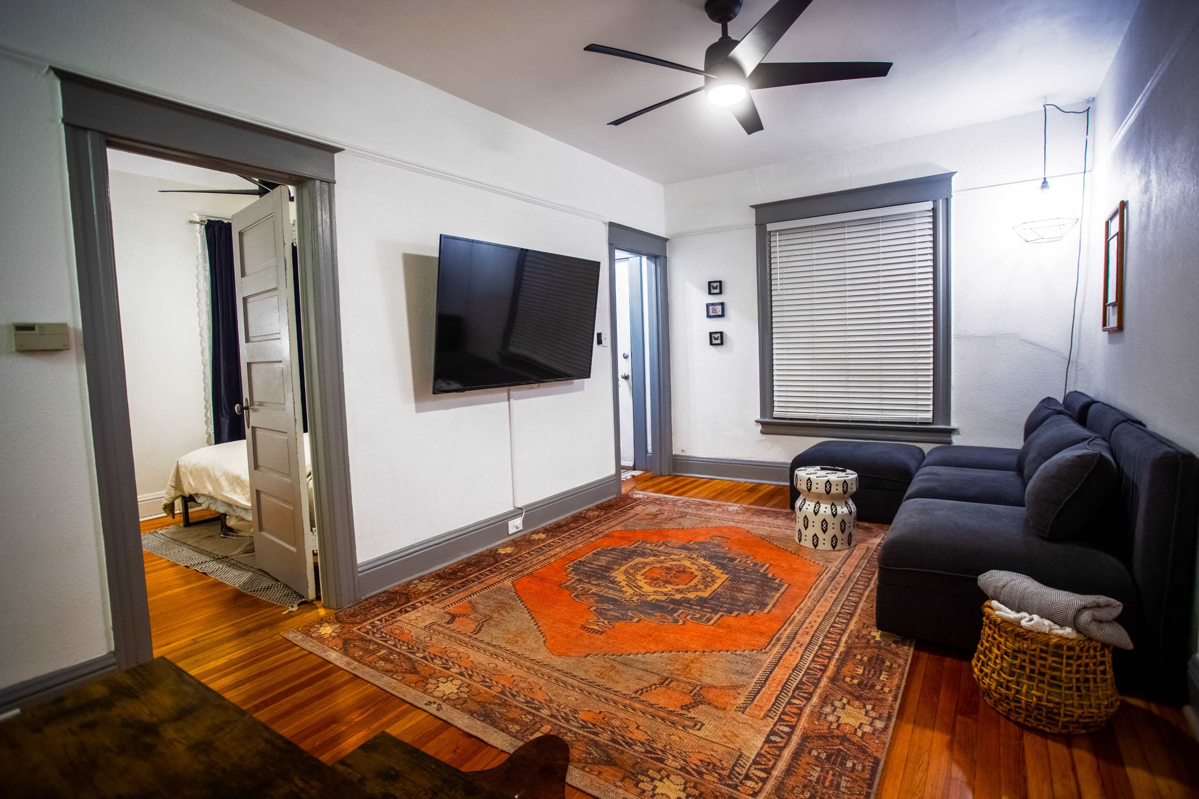 A living room with a dark couch, a large television mounted on the wall, and a colorful area rug on wooden flooring, leading to a doorway with a bed visible in the adjacent room.