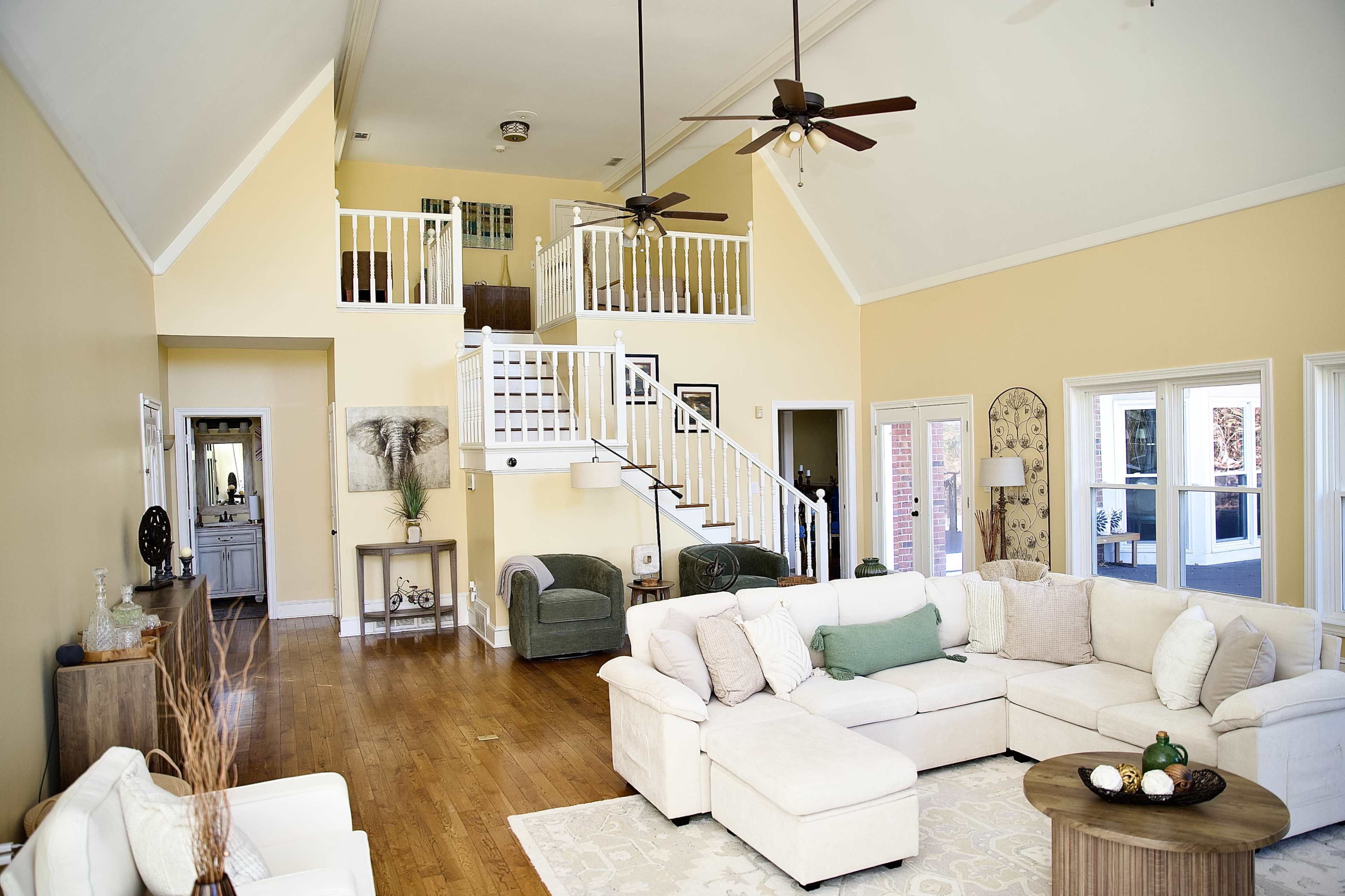 The image shows a spacious living room with a high ceiling, wooden floors, and a sectional sofa arranged around a round coffee table.