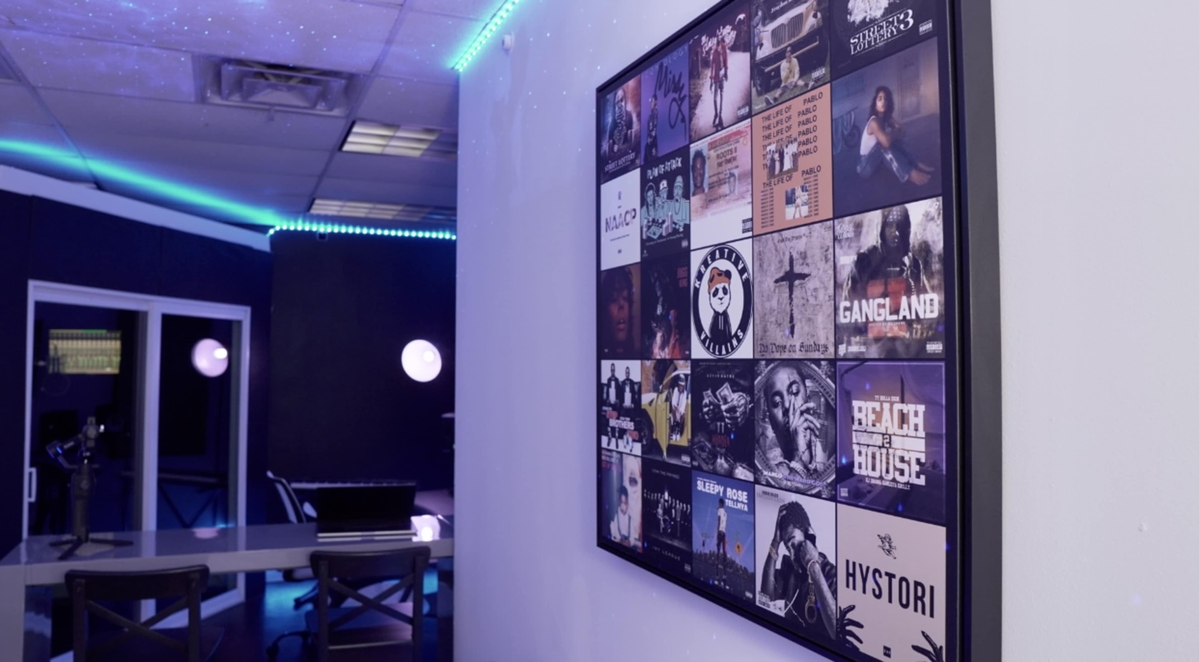 The image shows a wall-mounted collage of album covers beside a table and chairs with a computer setup in a dimly lit room.