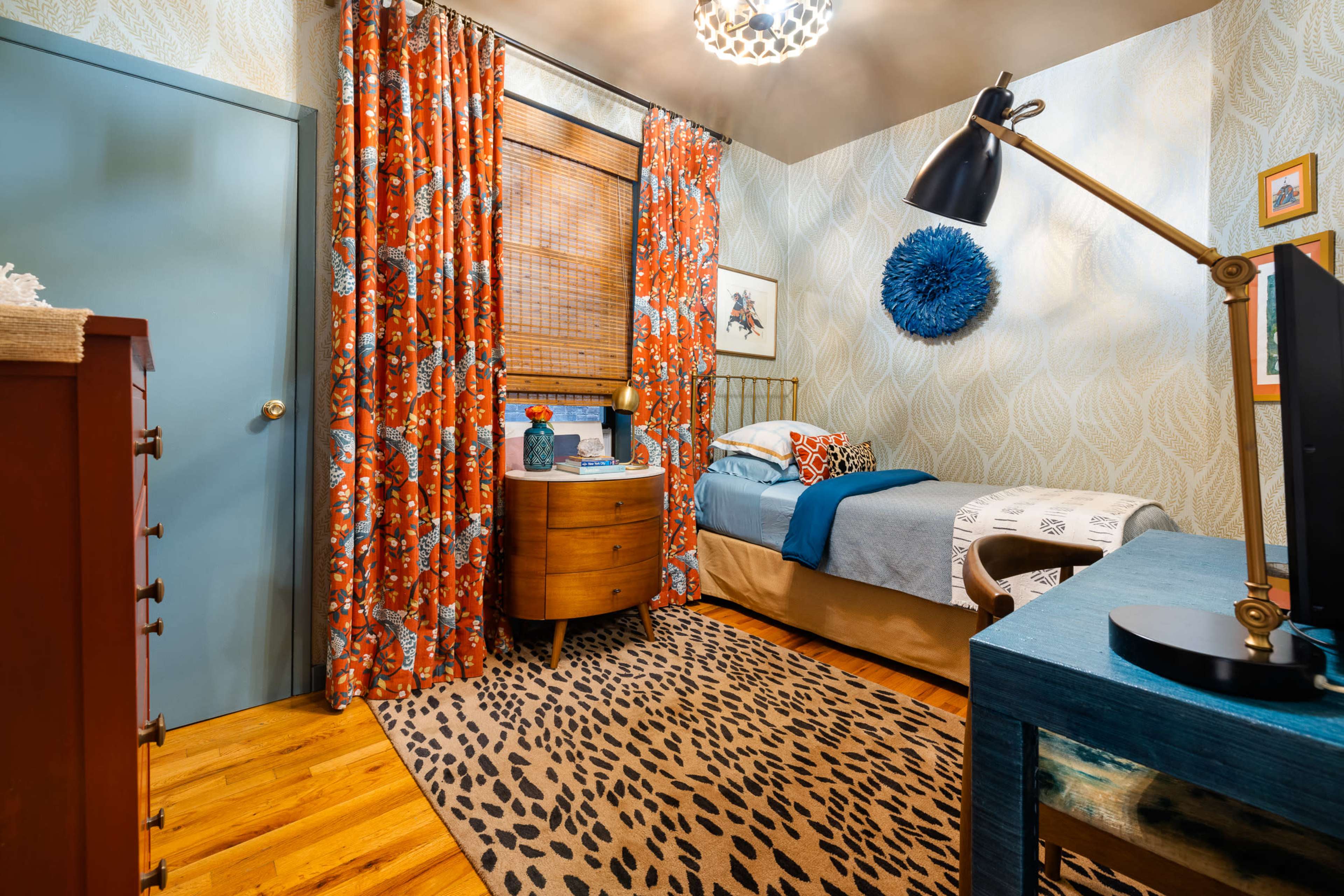 A small bedroom features a bed with blue bedding, orange patterned curtains, a wooden dresser, and a desk with a lamp, creating a cozy atmosphere with animal print decor.