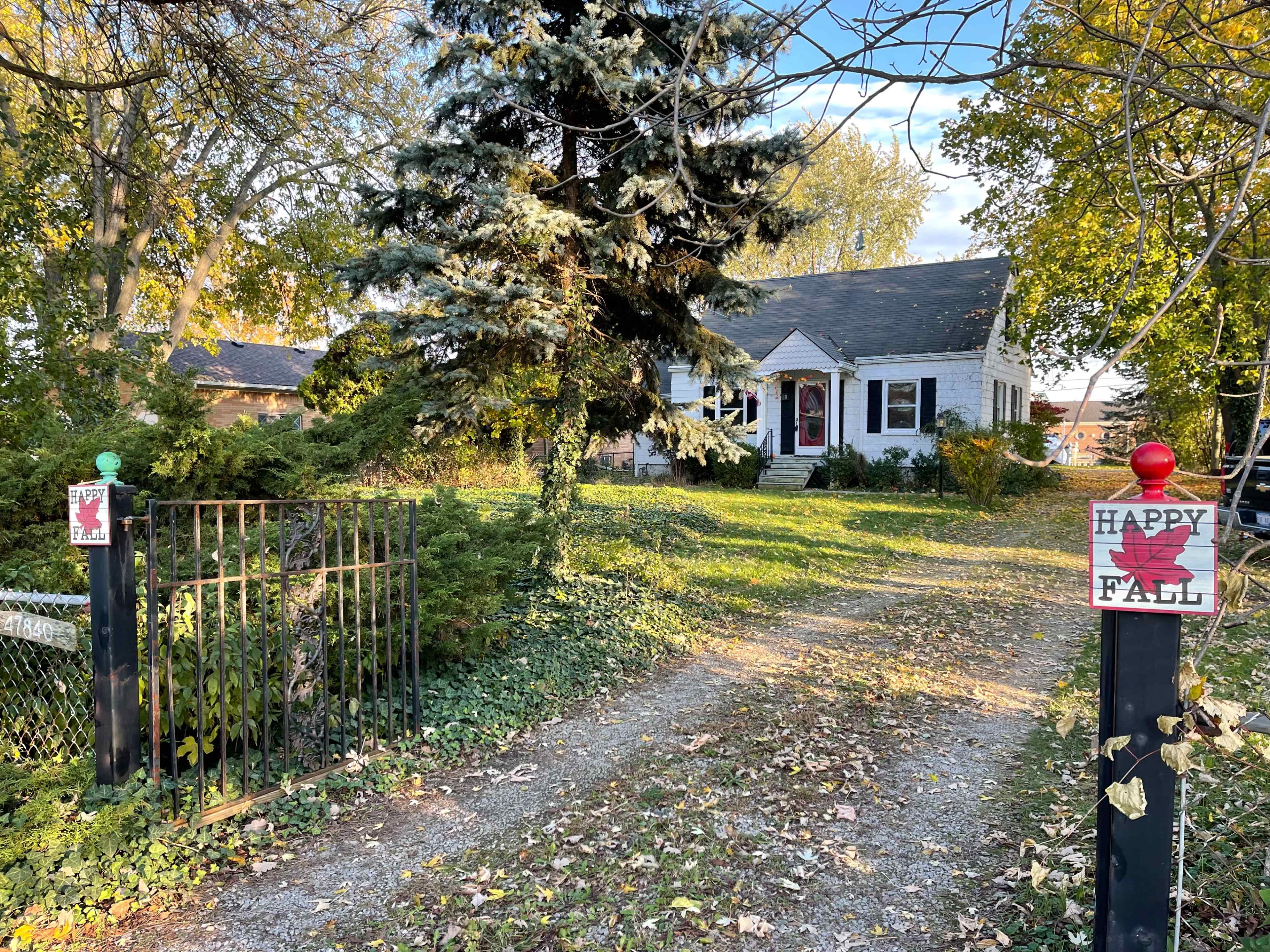 A gravel driveway leads to a white house surrounded by trees, with fall-themed signs at the entrance.
