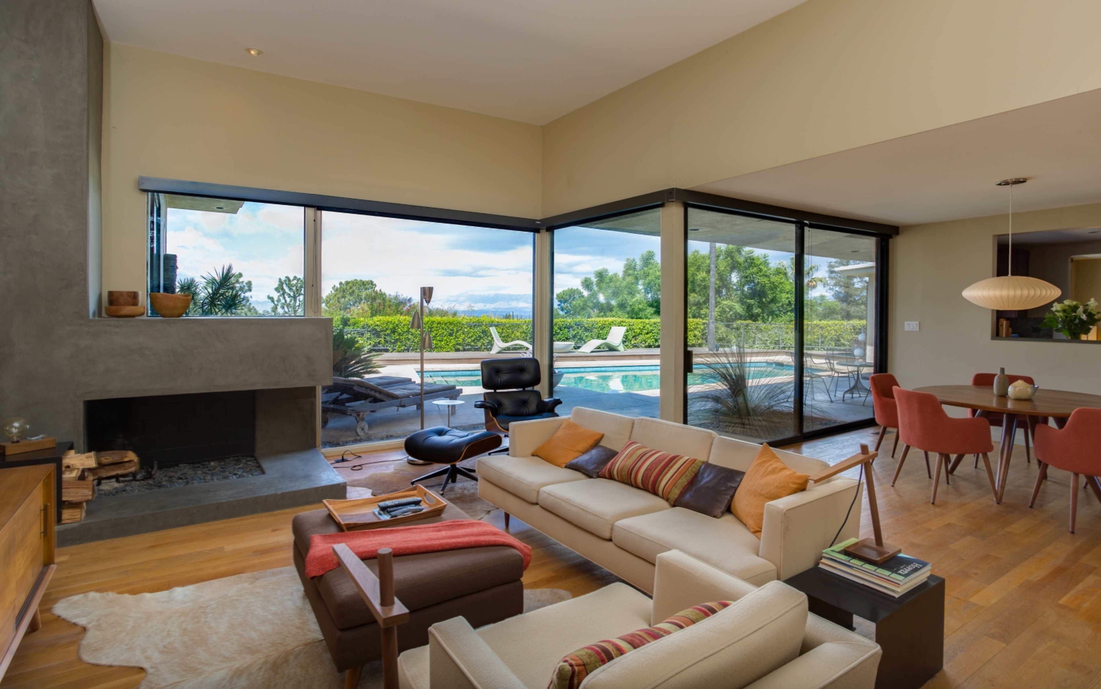 A modern living room features a large glass window overlooking a pool area and lush greenery, with a neutral-colored sofa and a simple dining area.