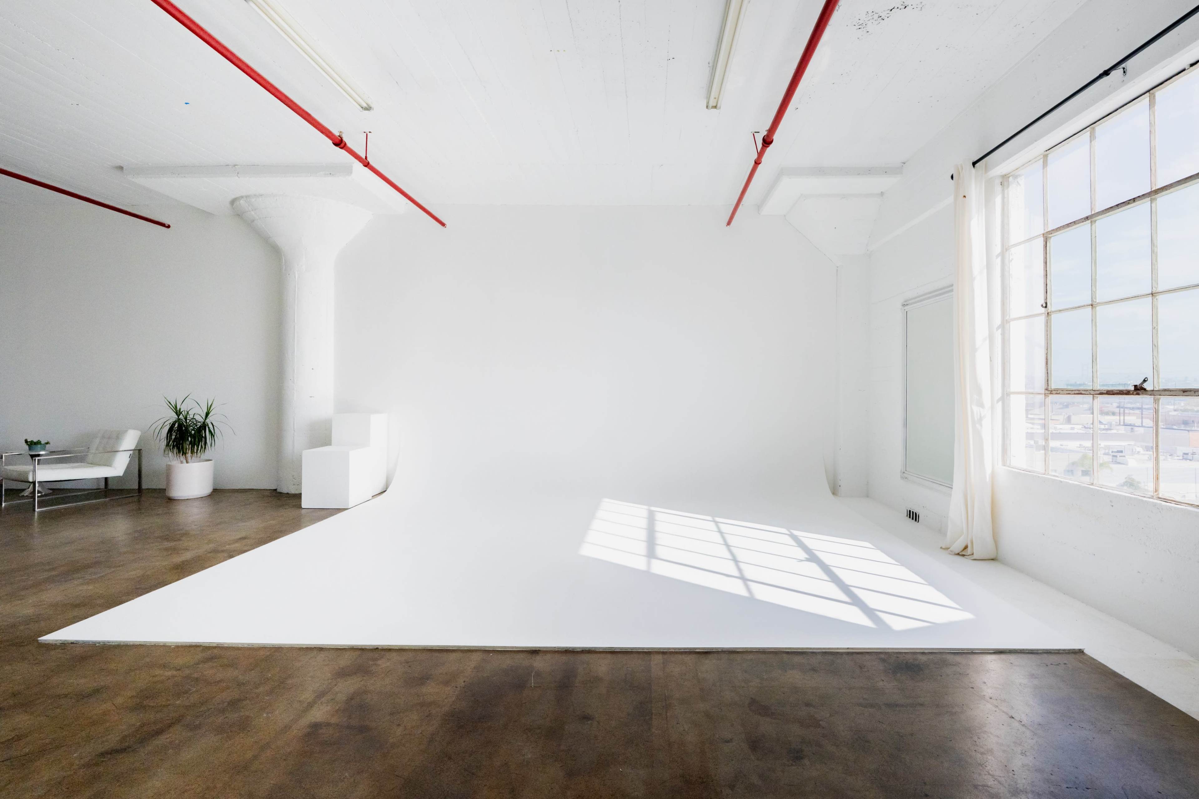The image shows a spacious, minimalistic studio with a white backdrop, large window, and industrial-style decor.
