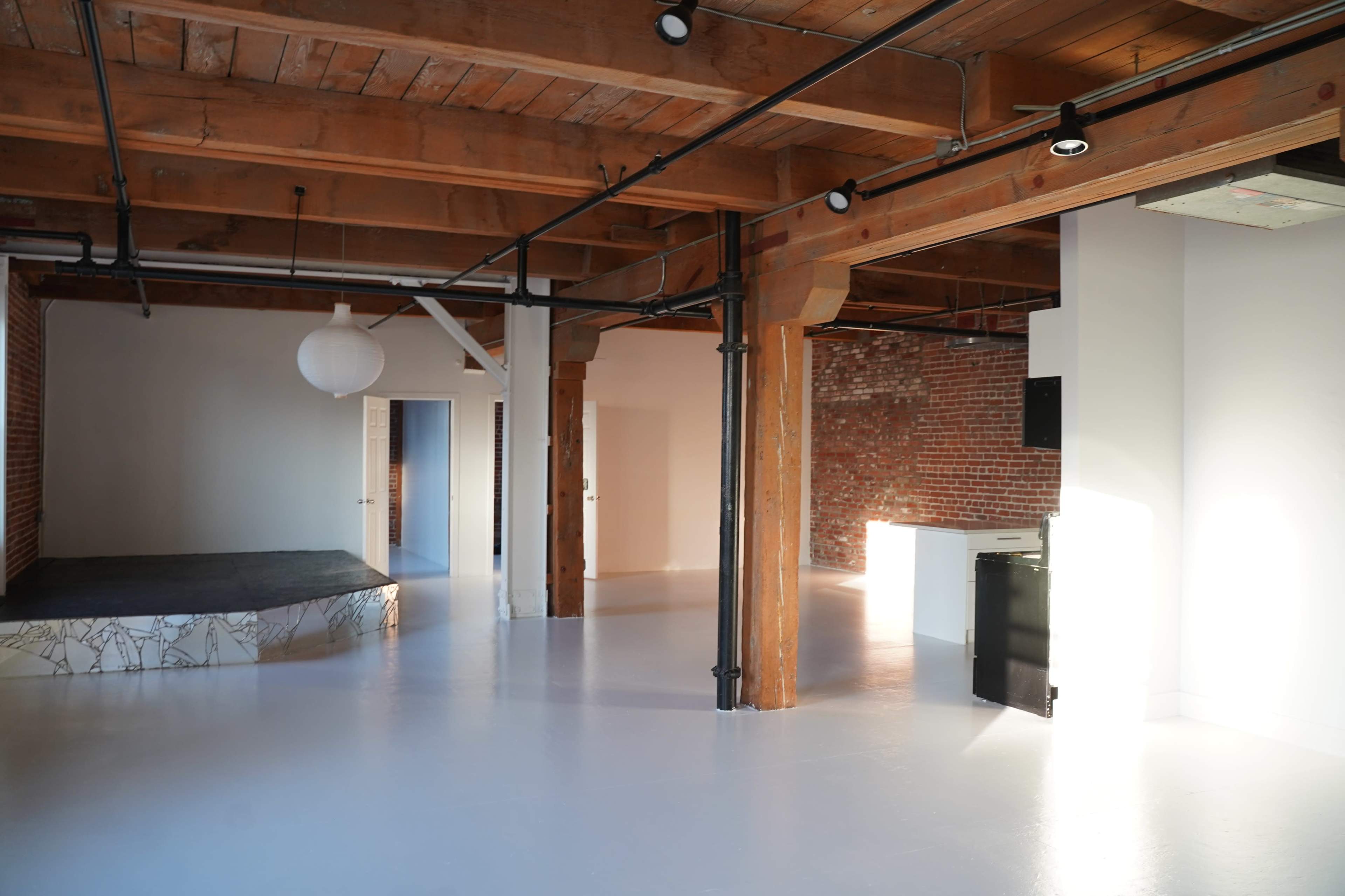 The image shows a spacious room with exposed wooden beams, a polished white floor, and a few distinct areas including a platform and a kitchenette.