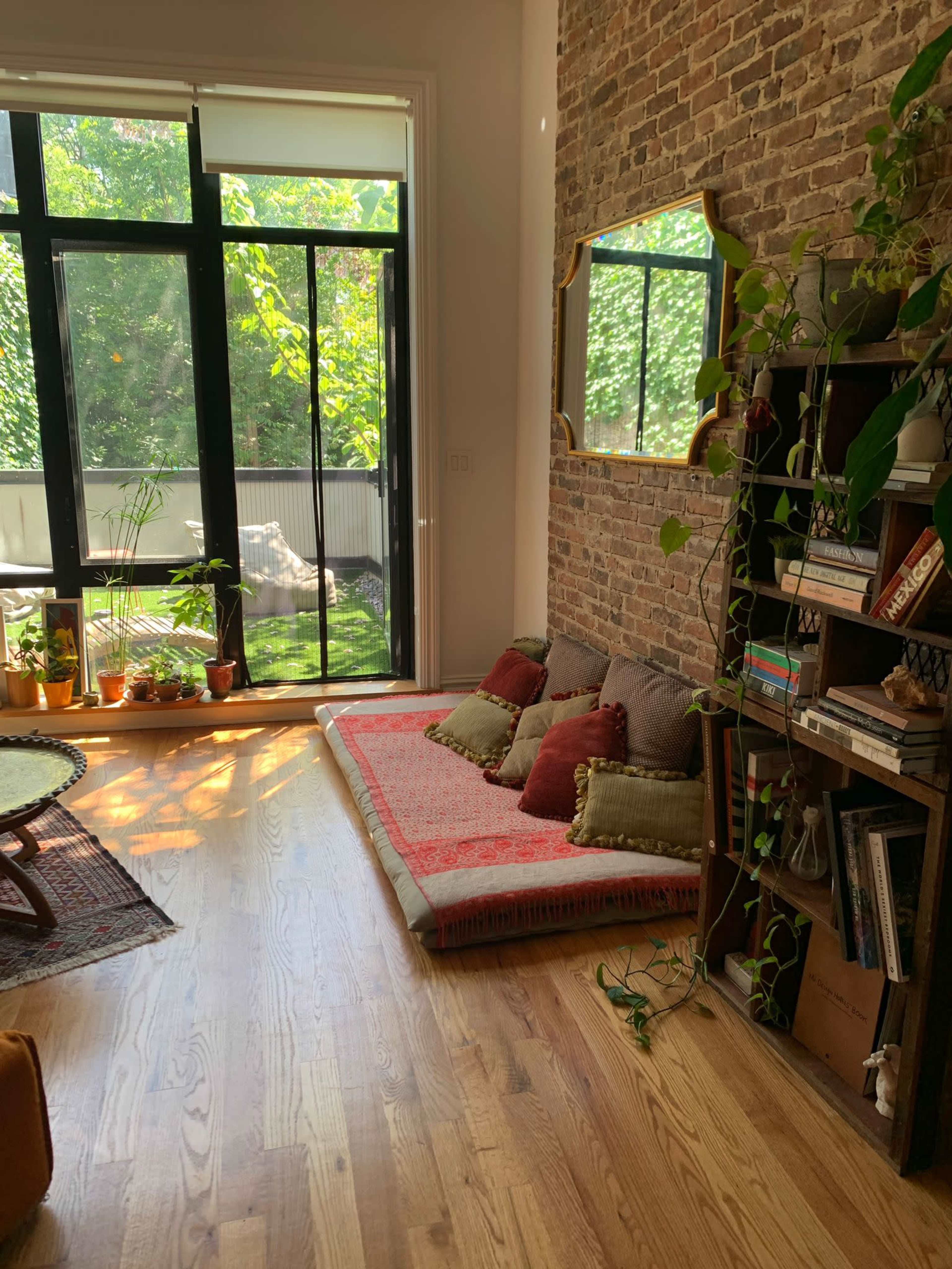 The image shows a cozy corner of a room with a brick wall, a cushioned seating area, a large window letting in natural light, and a bookshelf filled with plants and books.