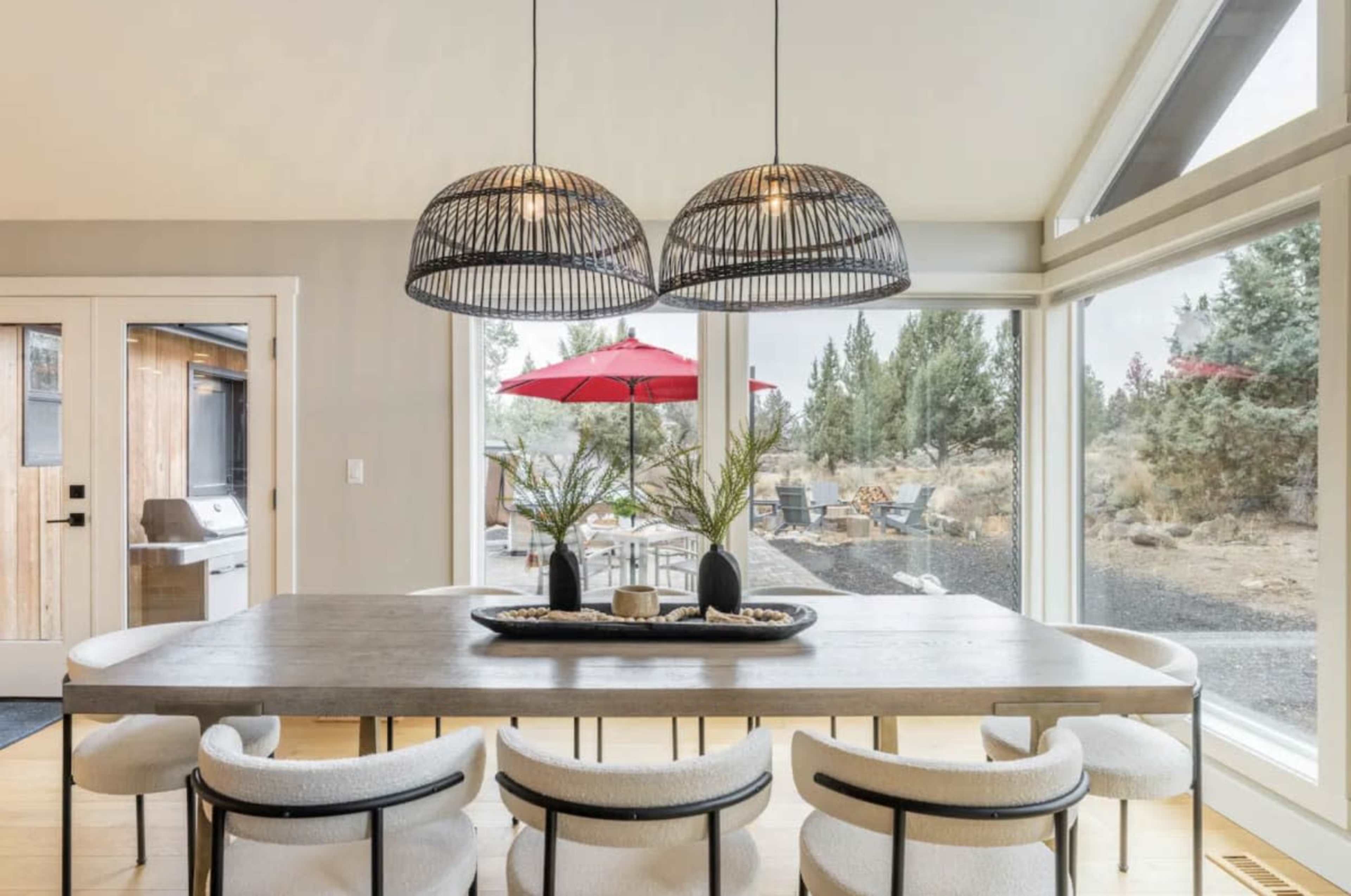 A modern dining area features a large wooden table surrounded by upholstered chairs, with two pendant lights hanging above and large windows overlooking an outdoor space with an umbrella and seating.