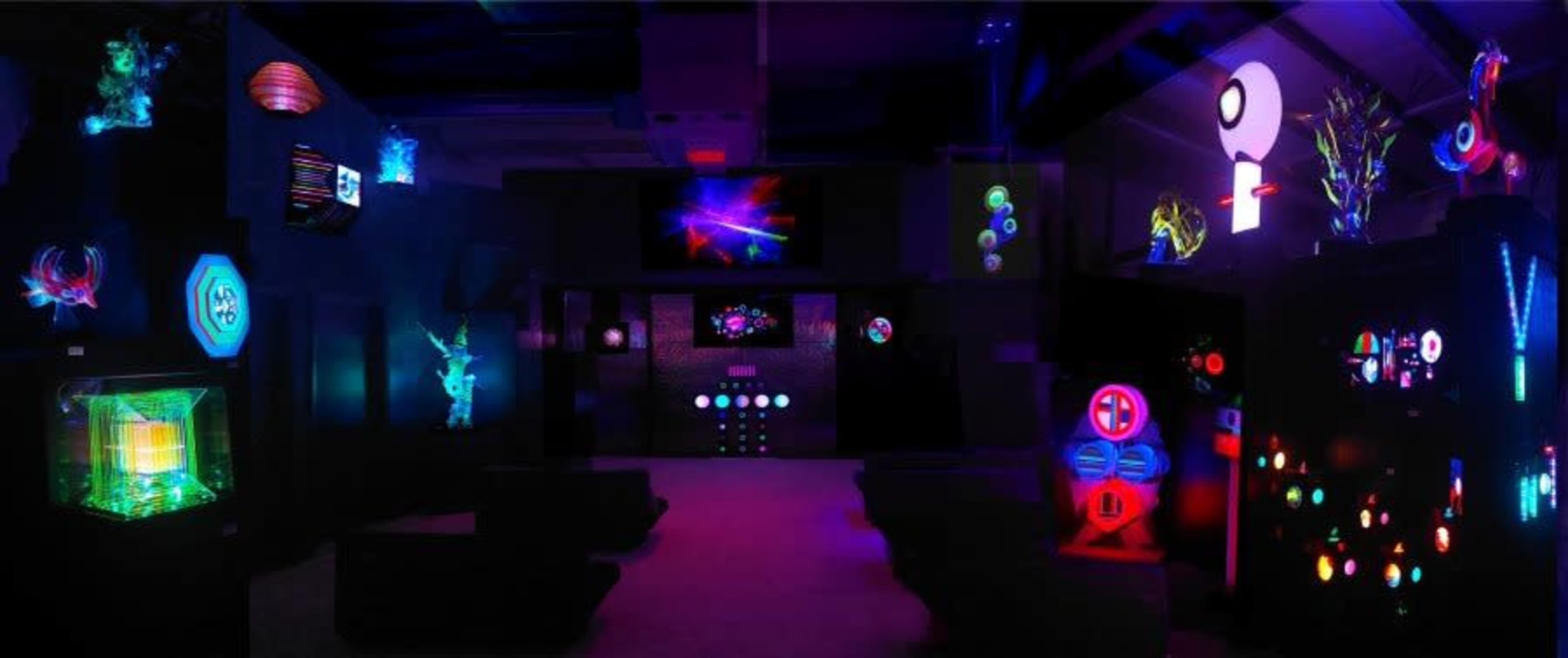 The image shows a darkened exhibition space filled with various illuminated sculptures and installations that feature vibrant colors and abstract designs.