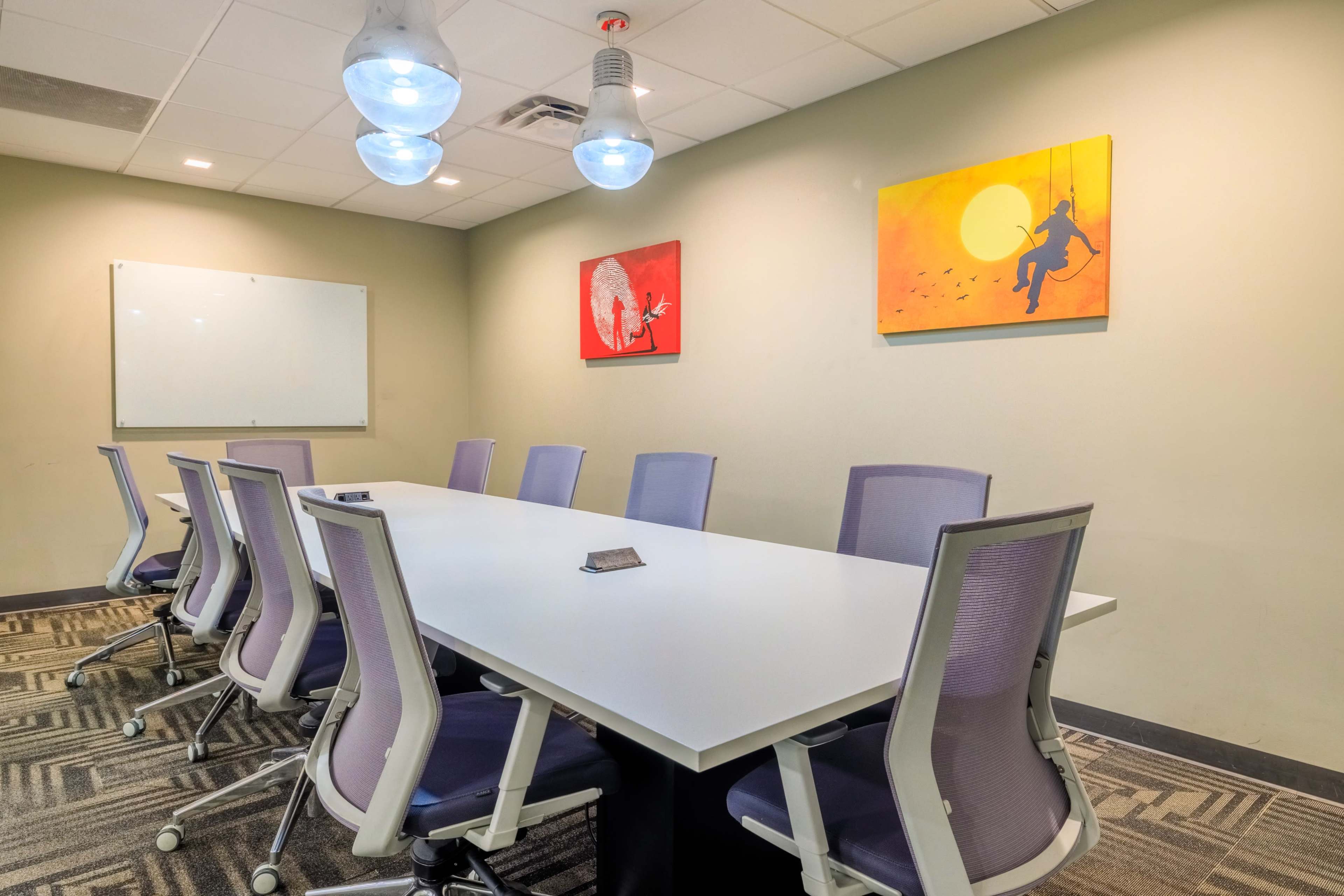 A modern conference room features a long white table surrounded by purple chairs, with two colorful paintings on the wall and a whiteboard at one end.