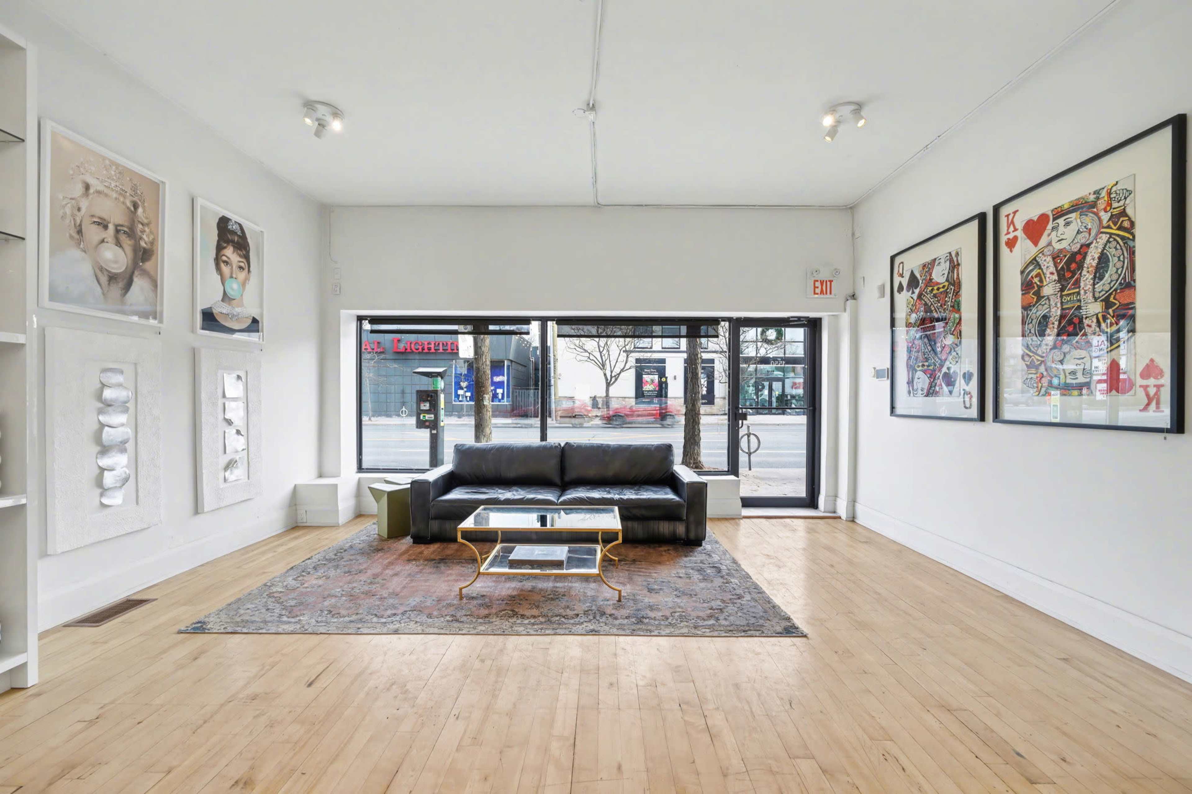 The image shows a modern gallery space with a black sofa, a glass coffee table, a large rug, and artwork displayed on the walls.