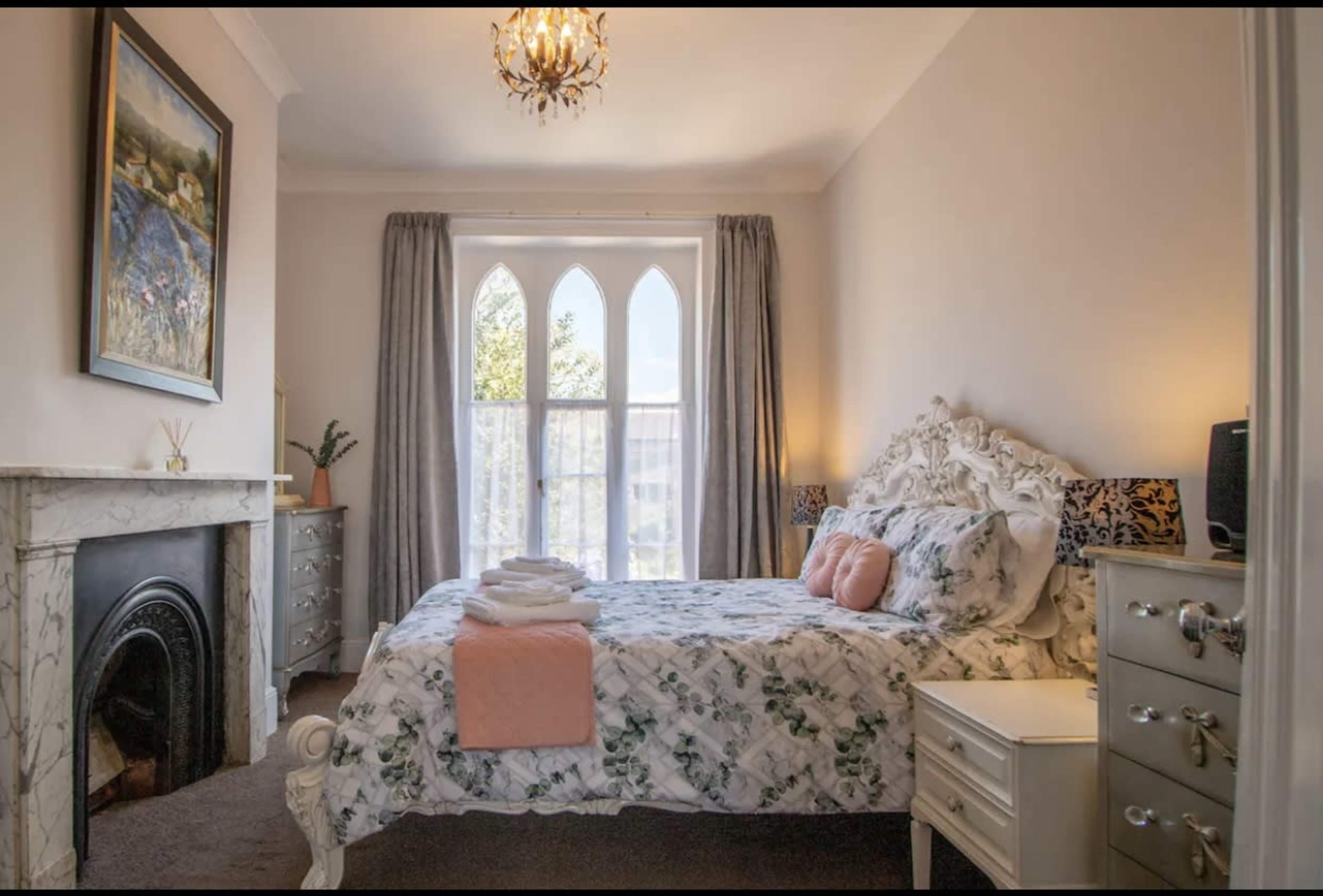 The image shows a neatly arranged bedroom featuring a bed with floral bedding, a marble fireplace, and large windows with arched tops.