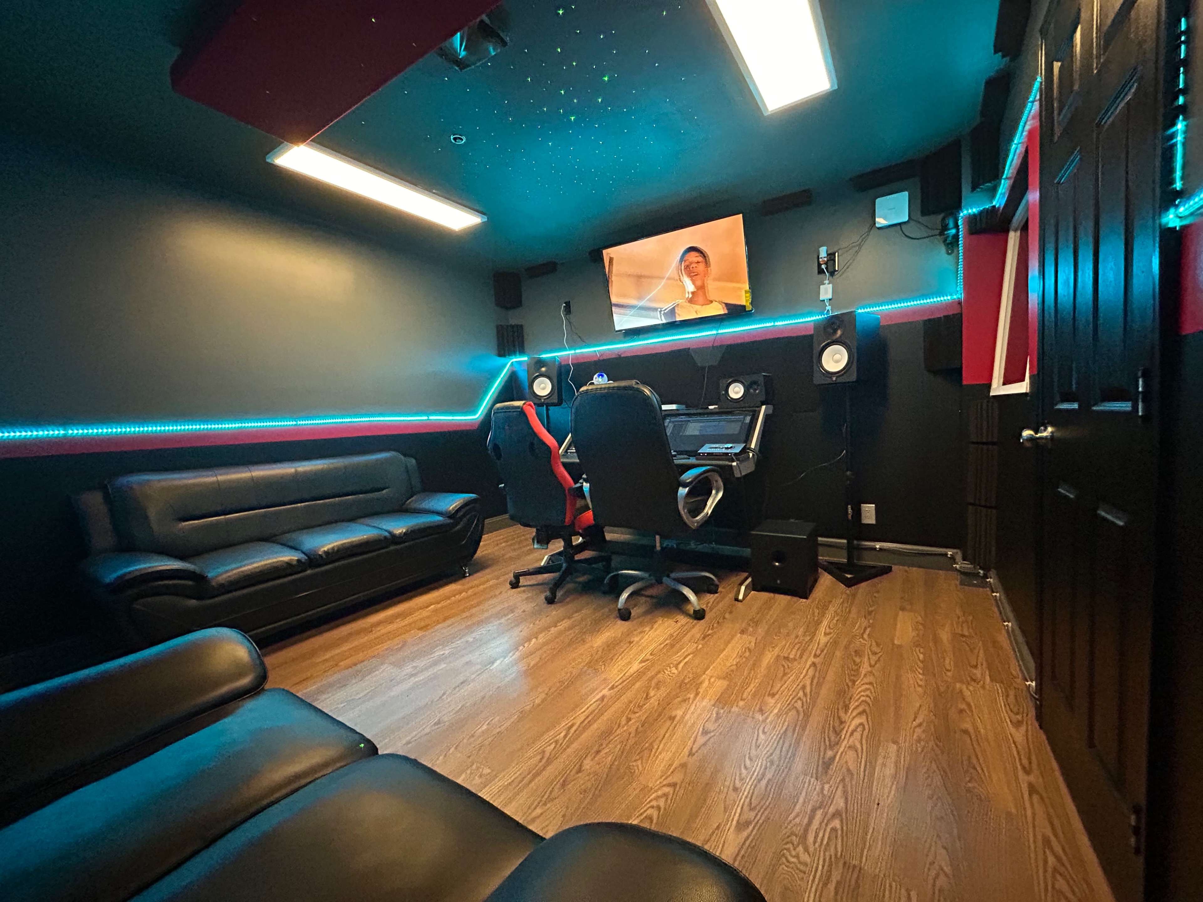 The image shows a music studio featuring a black sofa, a sound mixing desk with monitors, and a television mounted on the wall.