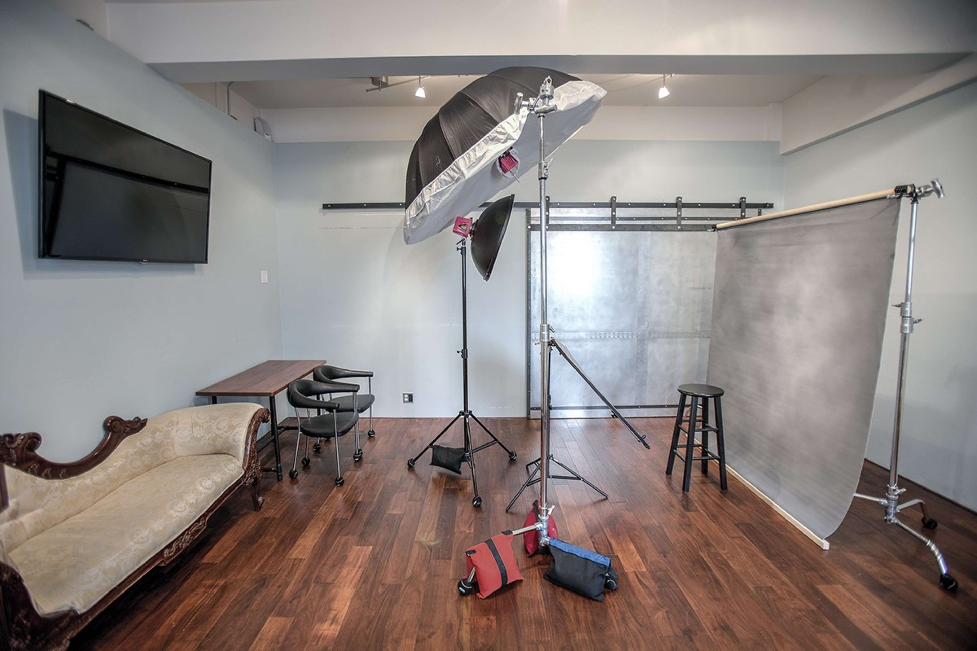 A photography studio featuring a lighting setup, a backdrop, a small table with chairs, and a chaise lounge against the wall.