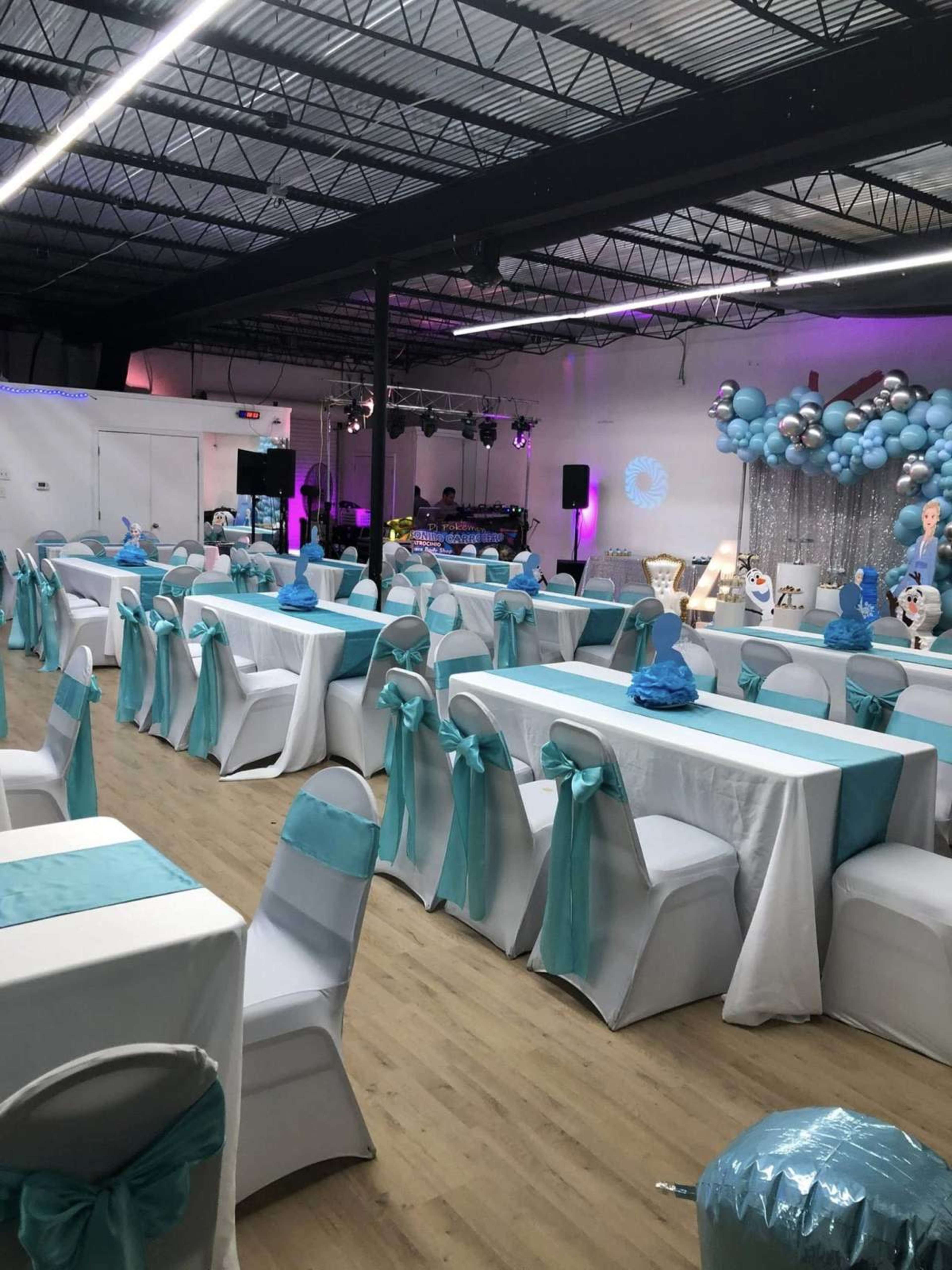 The image shows a decorated event space featuring white tables with turquoise accents, chairs adorned with matching bows, a DJ station, and balloon decorations in various shades of blue.