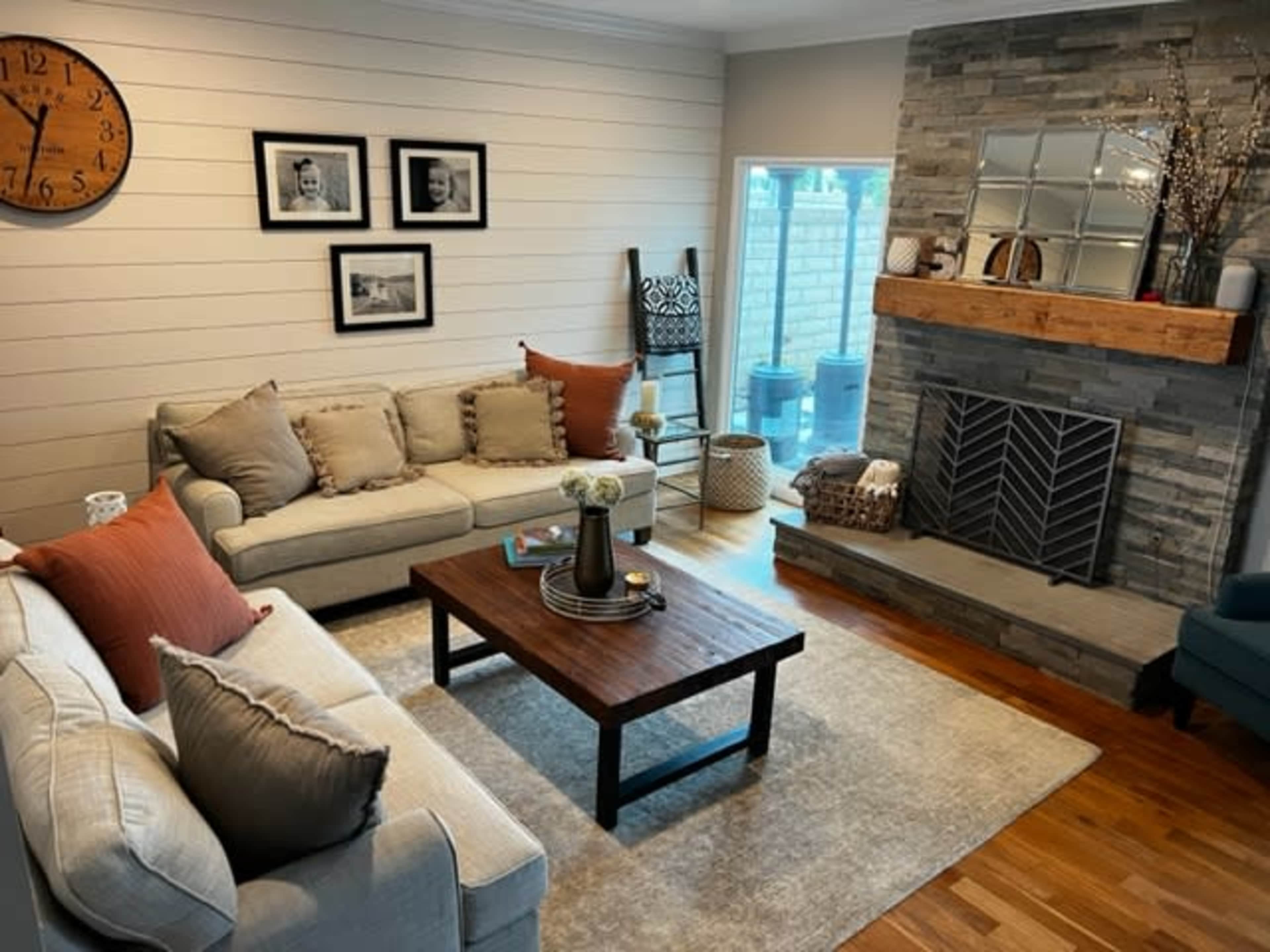 The image shows a modern living room featuring two sofas, a wooden coffee table, a stone fireplace with a wooden mantel, and large windows in the background.