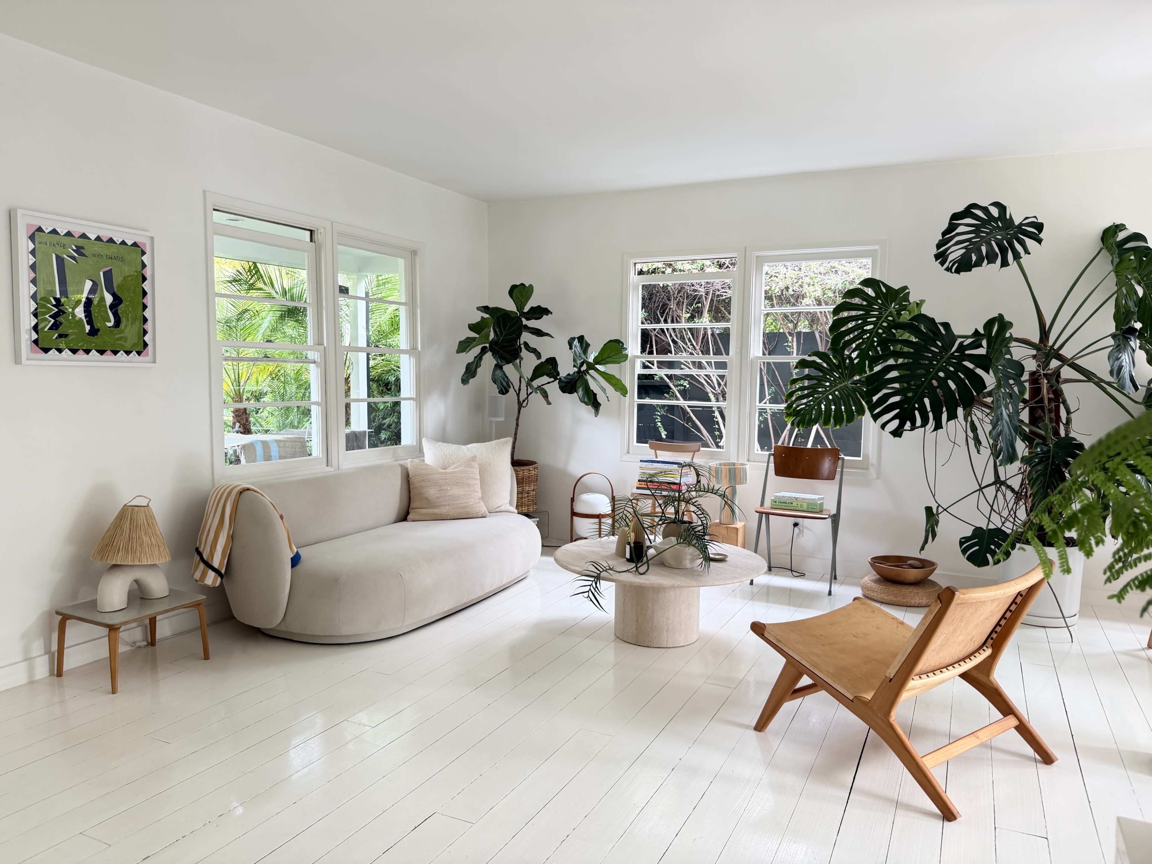 The image shows a bright, minimalist living room featuring a soft beige sofa, a circular coffee table, various potted plants, and a light wooden chair.