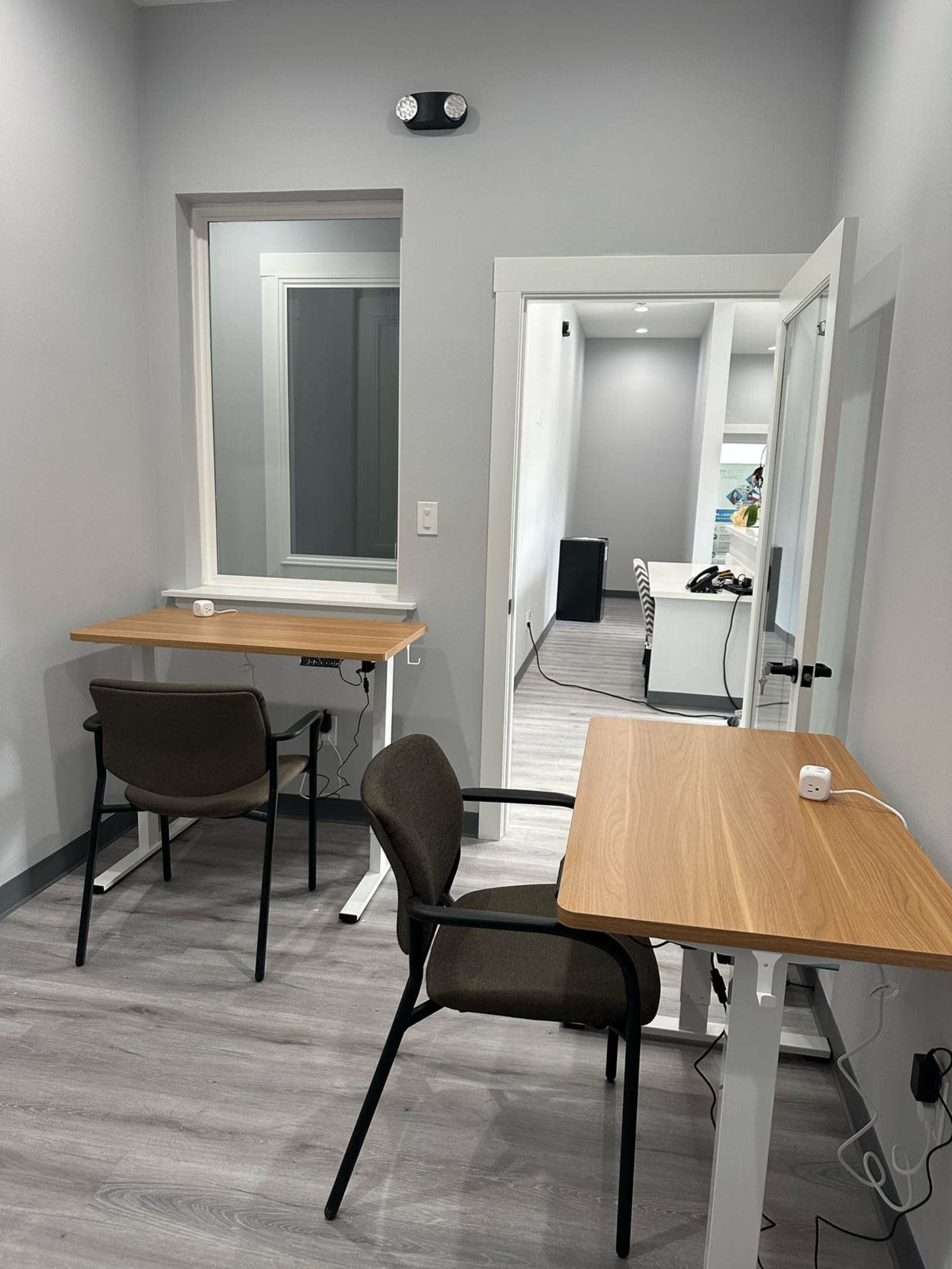 The image shows a small room with two wooden desks and chairs, one facing a window and the other positioned close to a doorway leading to another space.