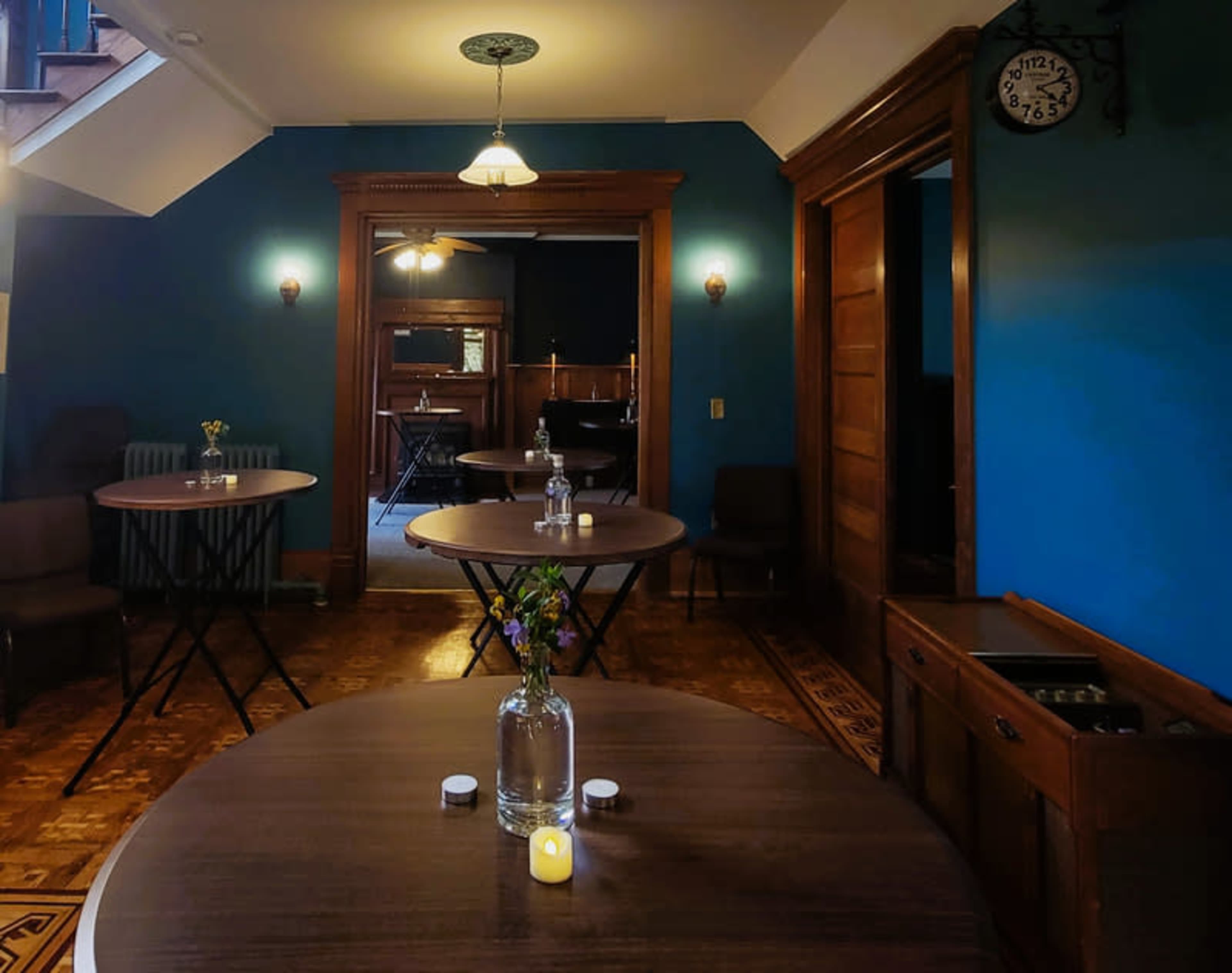 The image shows a room with two round tables set up, each with a vase of flowers, and a wall painted blue, featuring wooden doors and a clock.