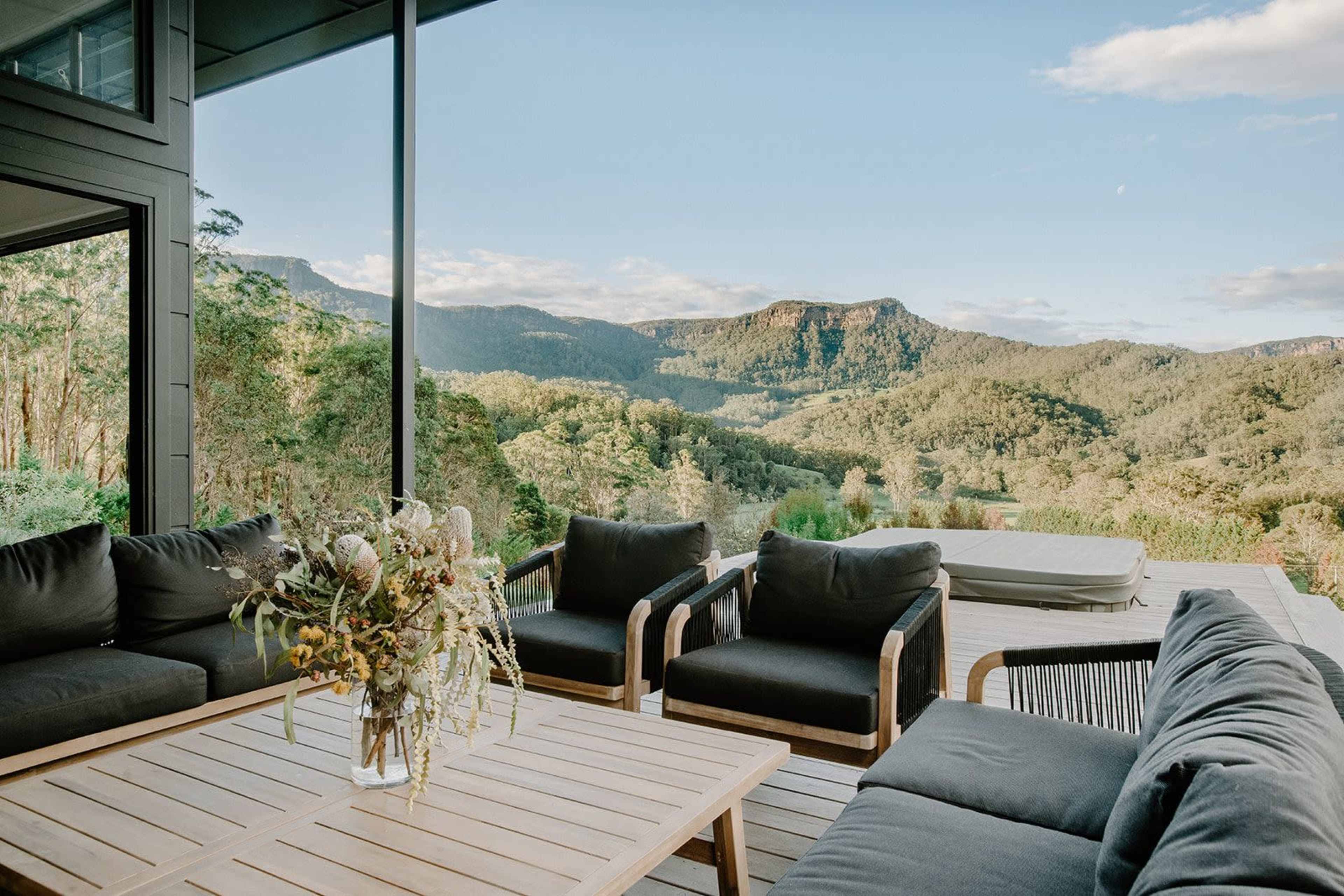 A modern outdoor living area features comfortable seating and a wooden coffee table, overlooking a lush mountain landscape through large glass windows.