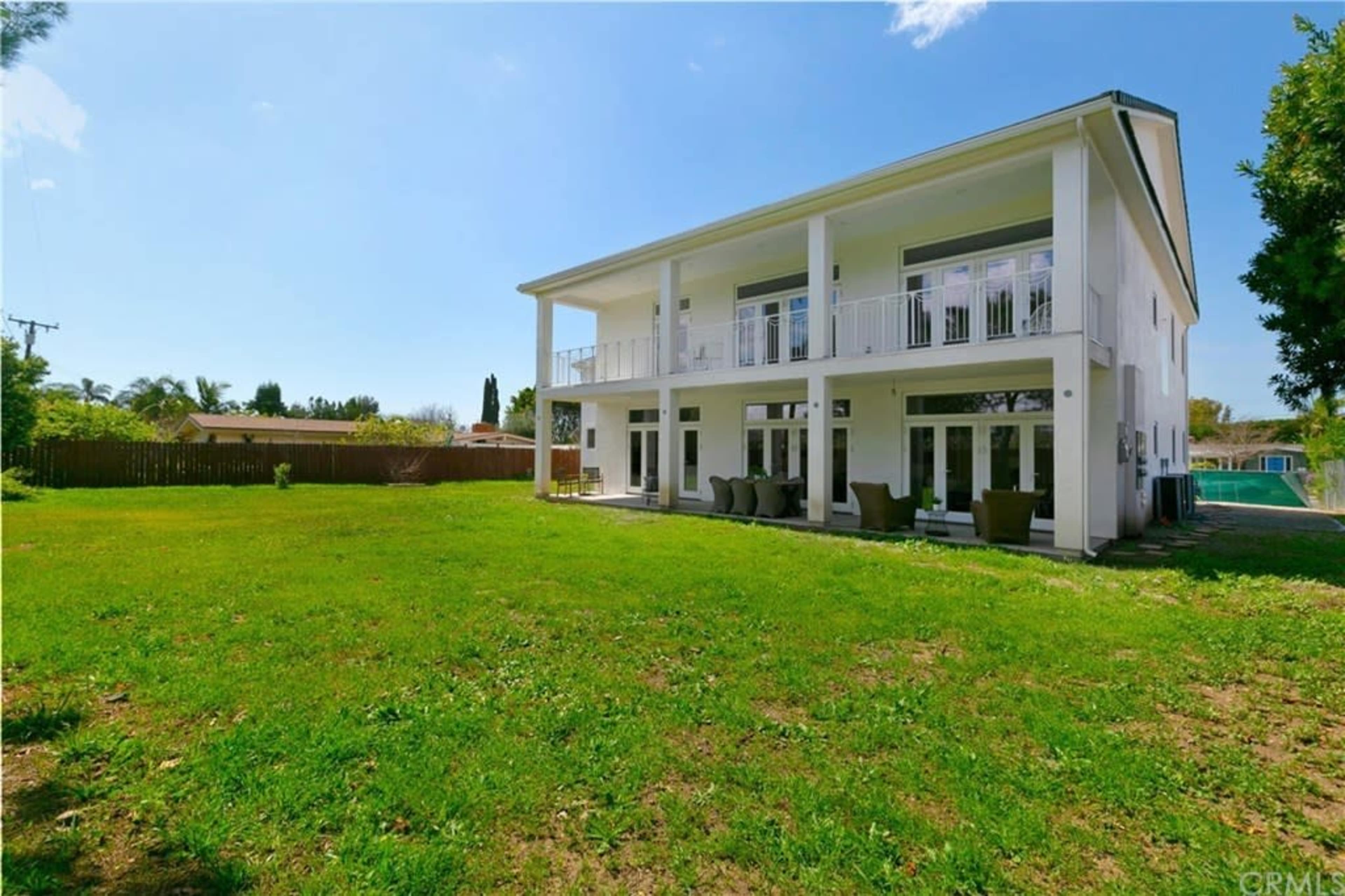 The image shows a two-story modern house with a large green lawn and a patio at the back, surrounded by trees and a clear blue sky.