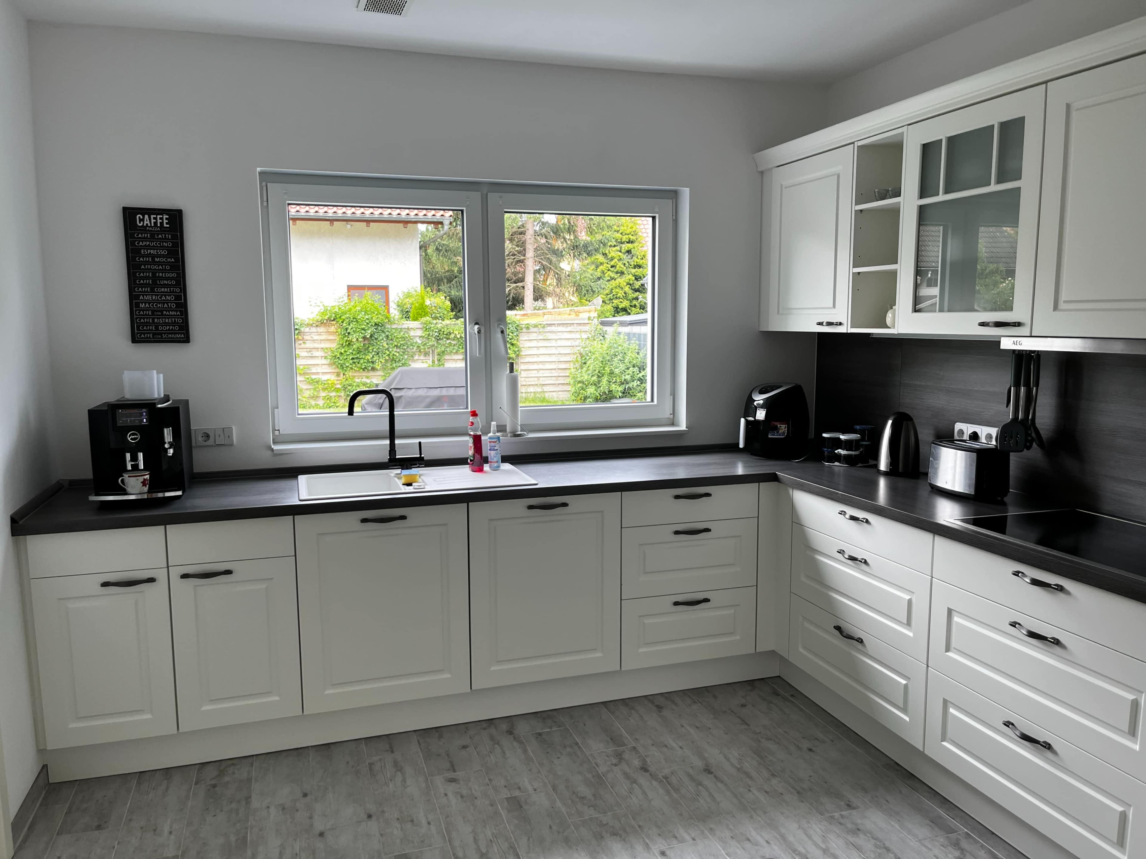 A modern kitchen features white cabinetry, a black countertop, and a window overlooking a garden.