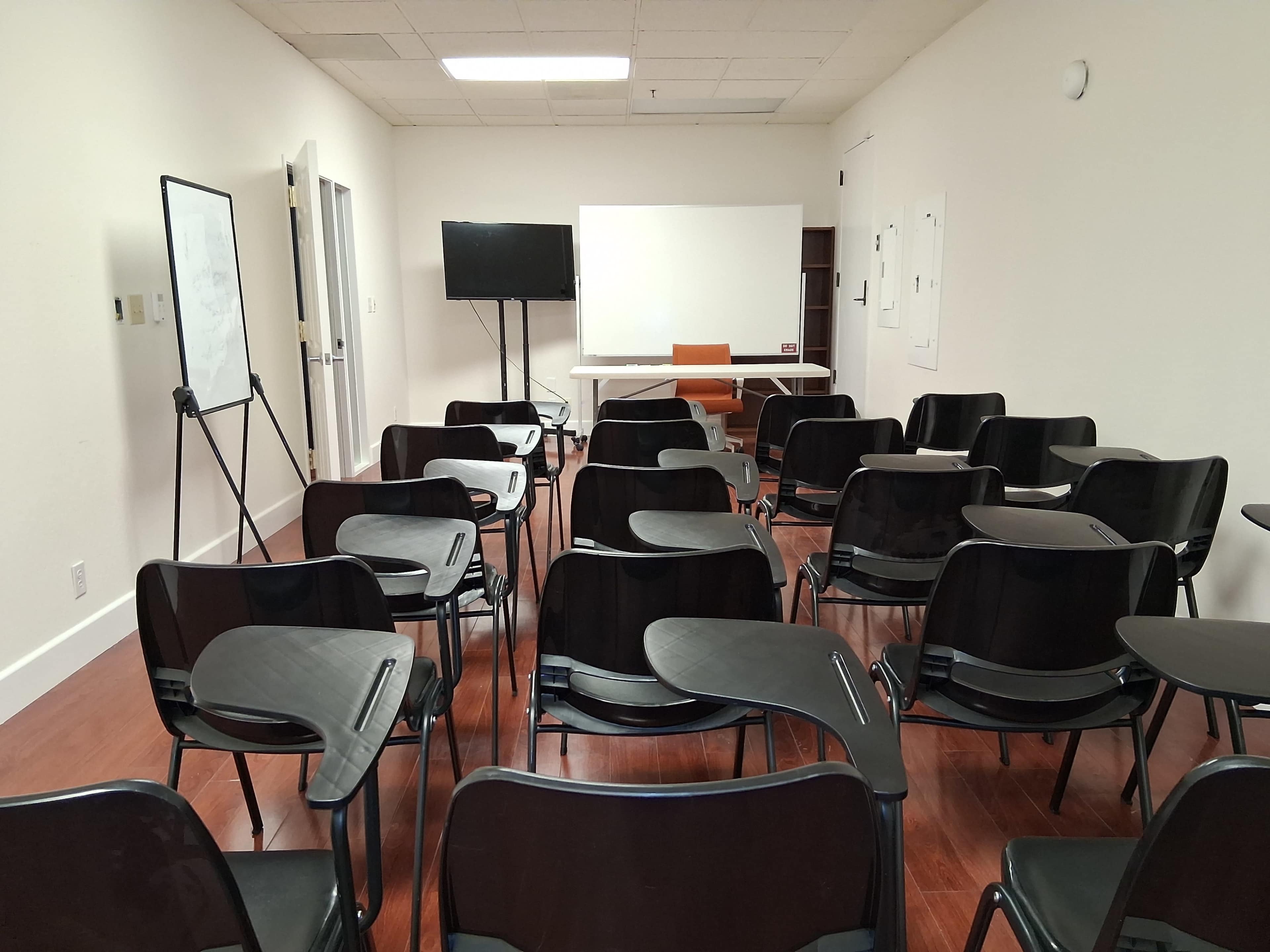 The image shows a classroom with rows of black chairs, a whiteboard on an easel, a TV on a stand, and a table with an orange chair in front of a blank white wall.