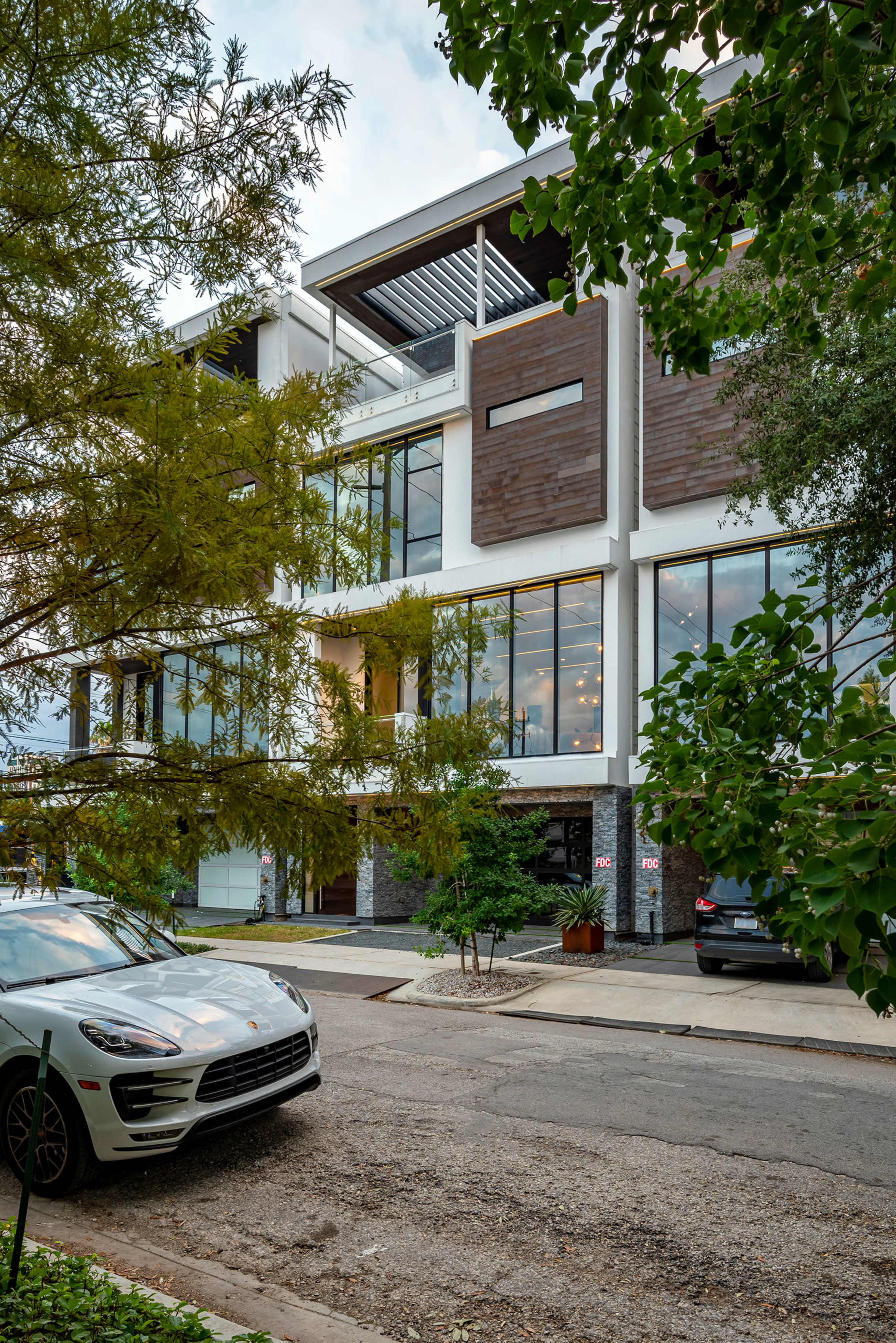 A modern multi-level building with large windows is seen from the street, surrounded by trees and parked cars.