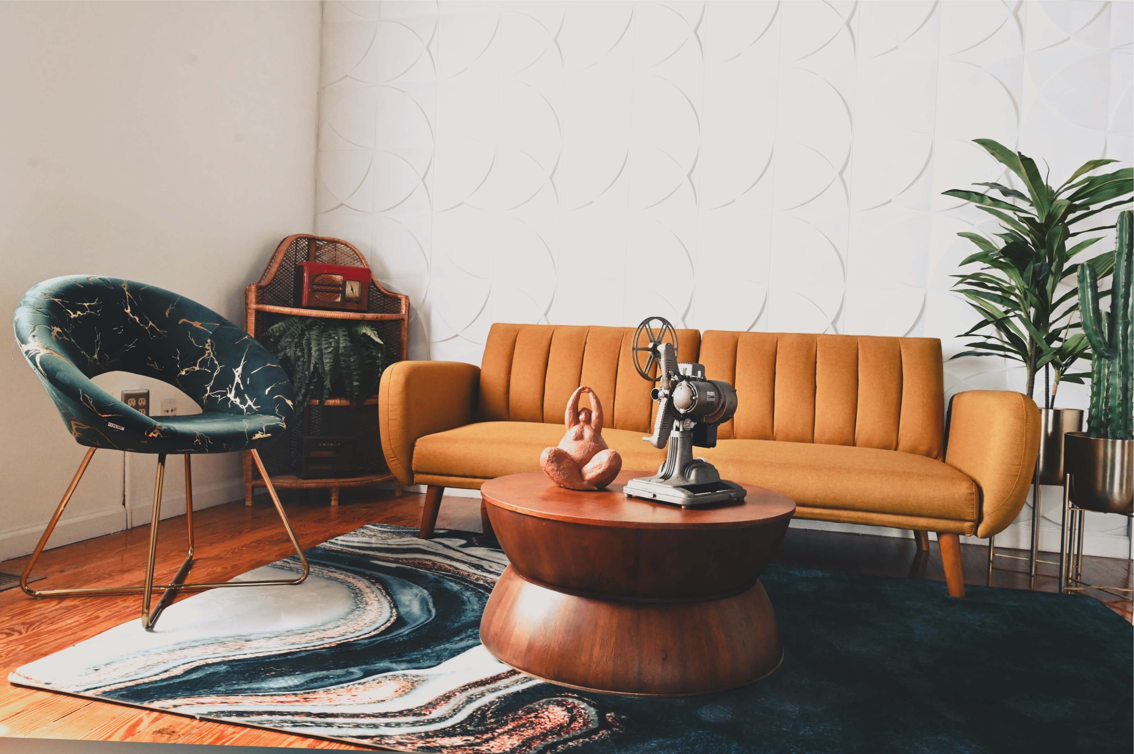 A stylish living room features a mustard yellow sofa, a unique green chair, a rounded wooden coffee table, and decorative plants against a textured white wall.