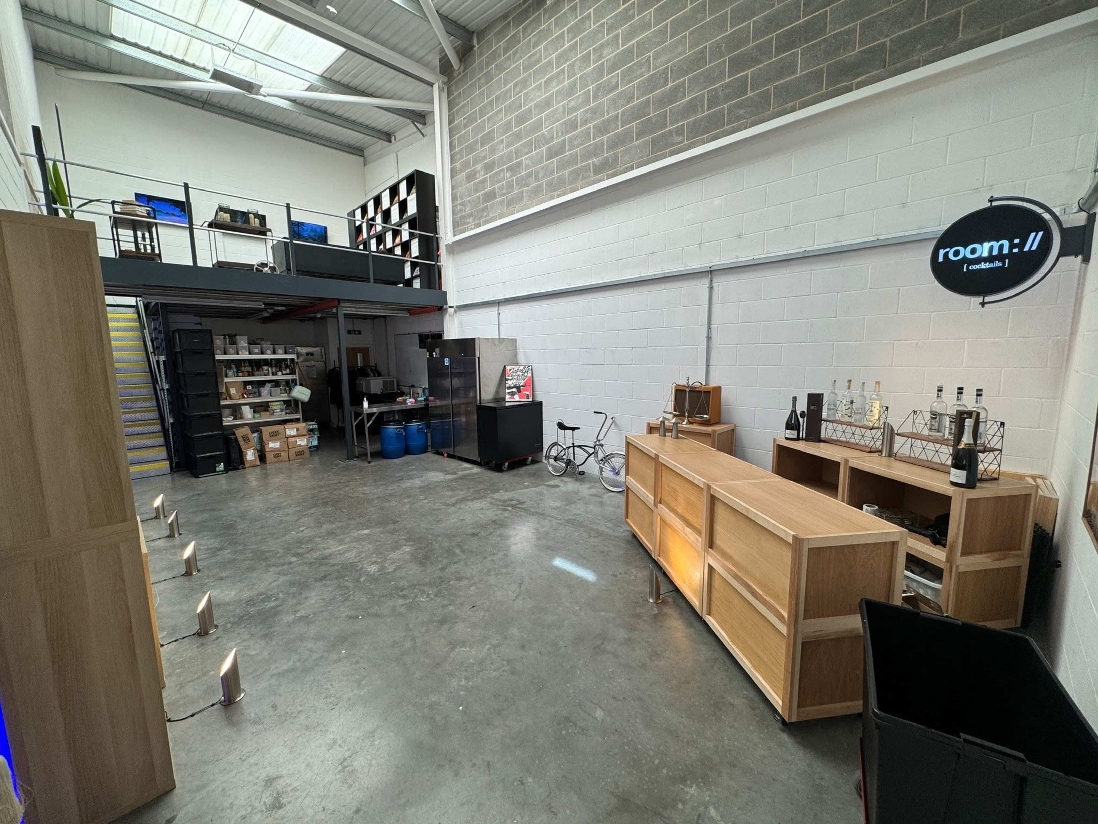 The image shows a spacious industrial interior with a wooden counter, shelving units, storage boxes, a bicycle, and a visible staircase leading to an upper level.