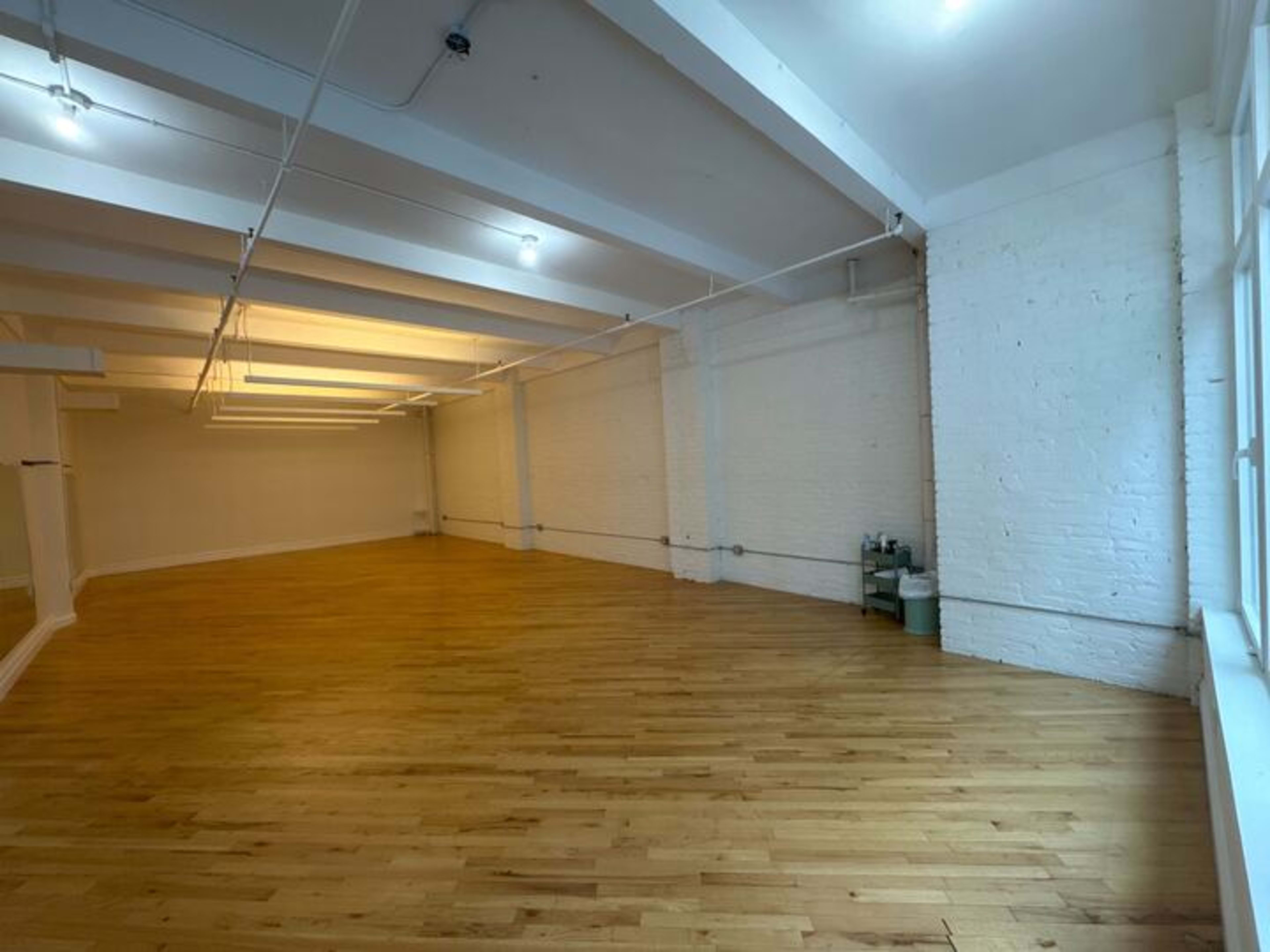 The image shows a spacious, empty room with wooden flooring and white walls, illuminated by overhead lights.