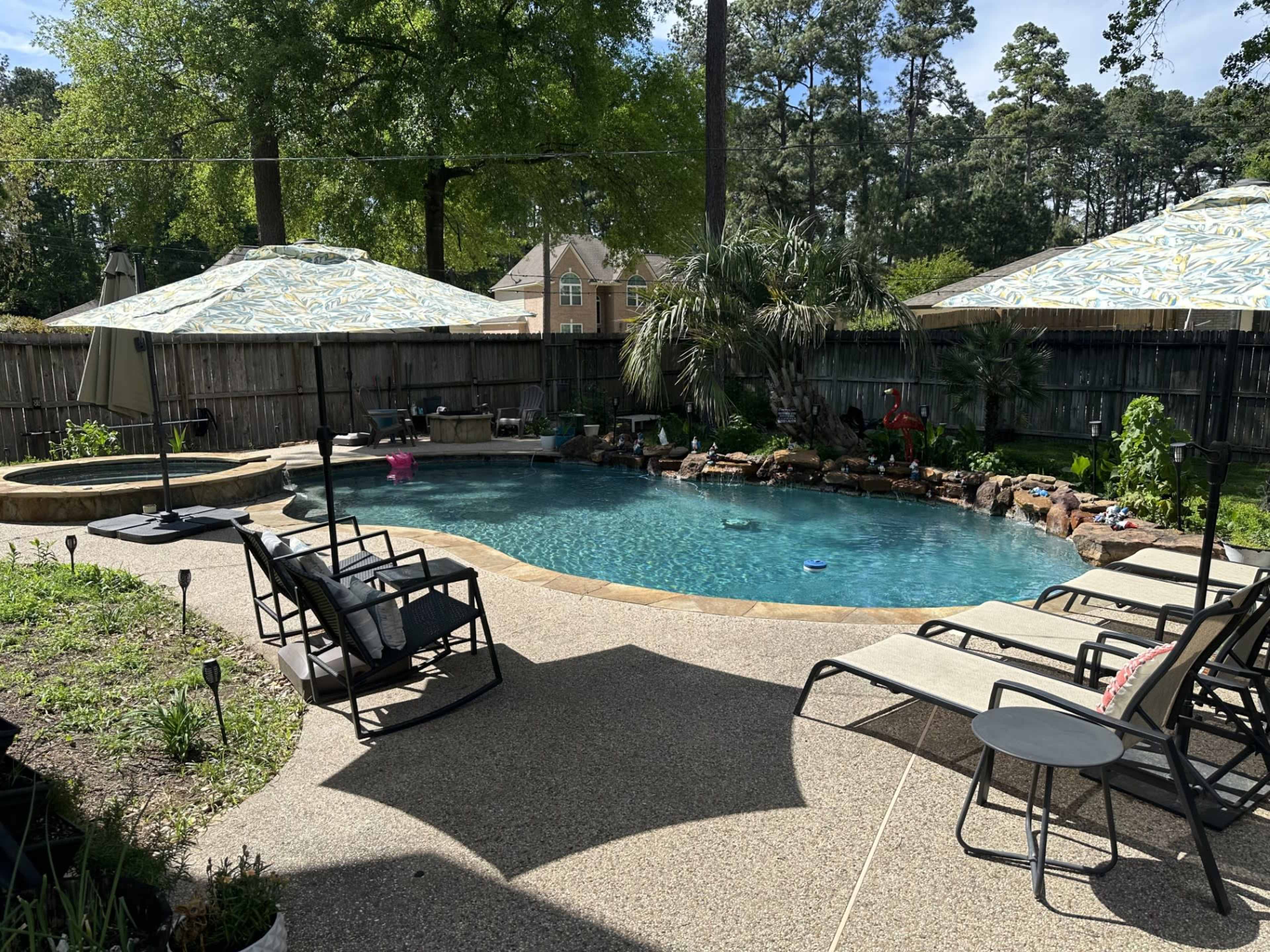 A backyard pool area features a clear blue swimming pool surrounded by lounge chairs and shaded by umbrellas.